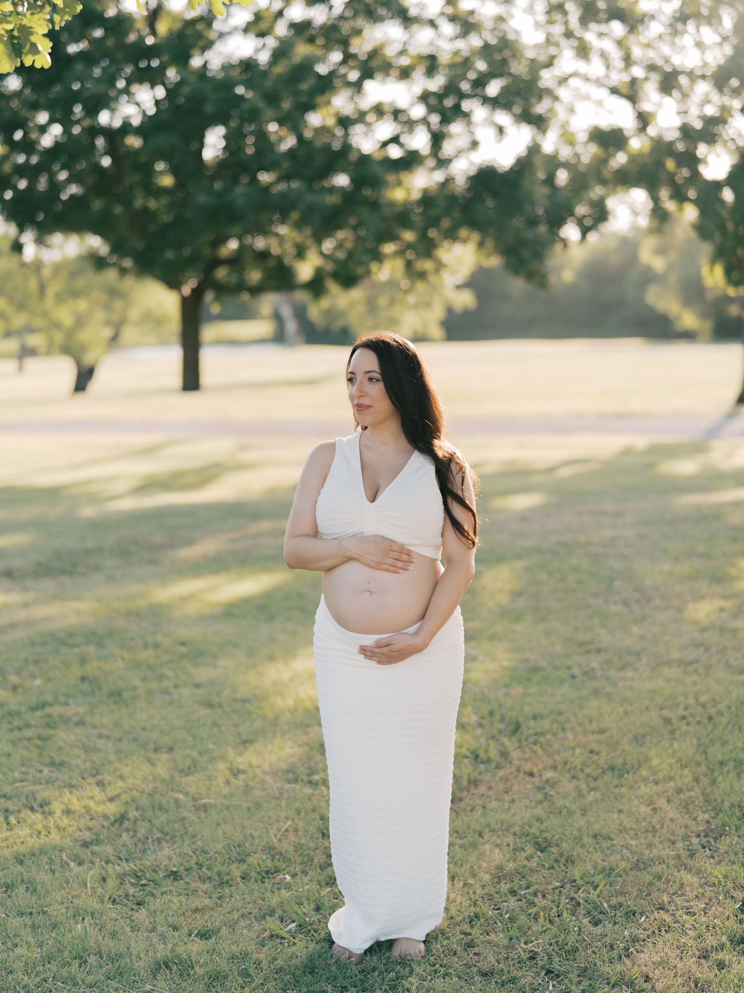 Pregnant woman in white maxi dress cradling baby bump in golden hour park setting.