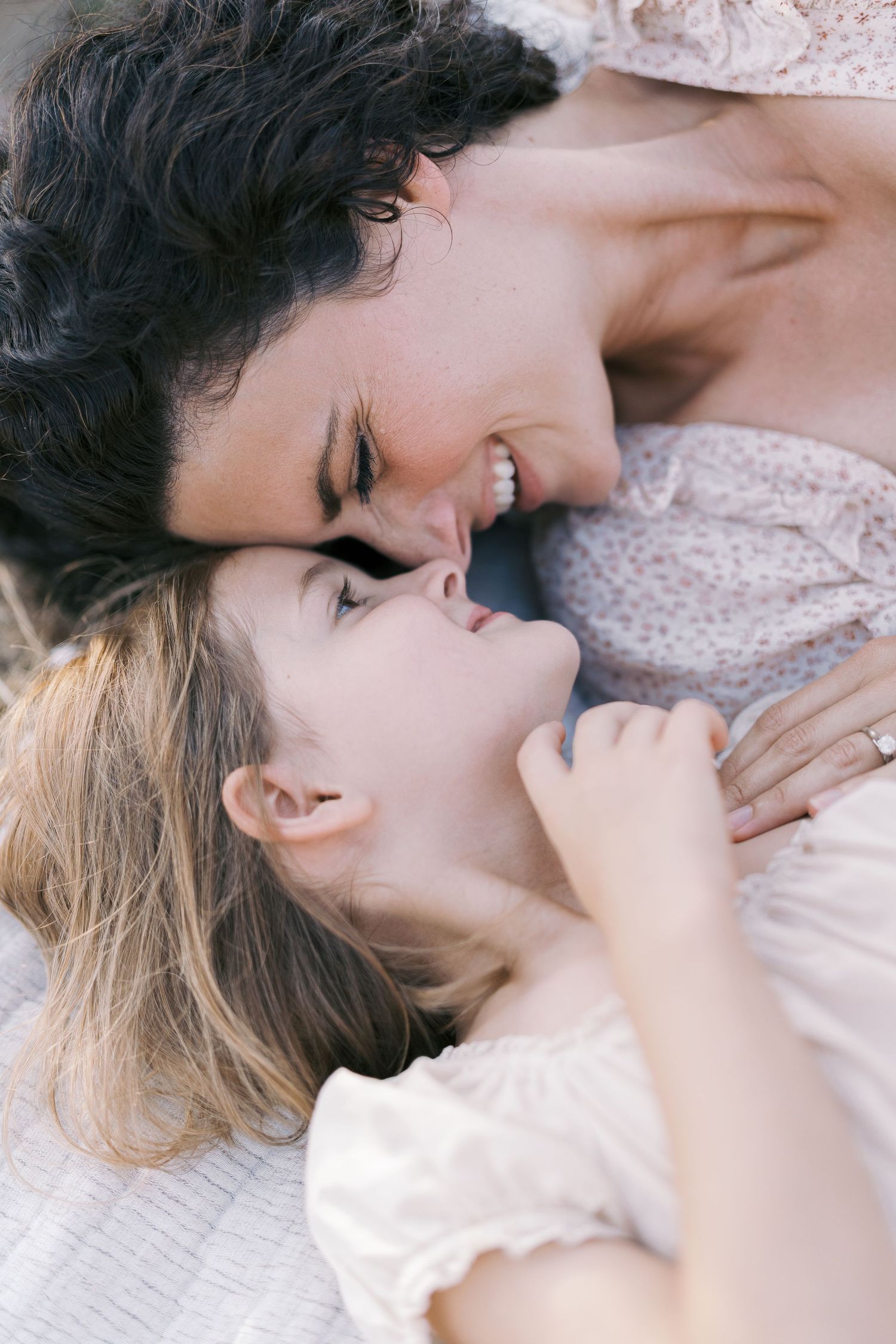 A mother and young daughter share a tender nose-to-nose moment while lying down, smiling warmly at each other.