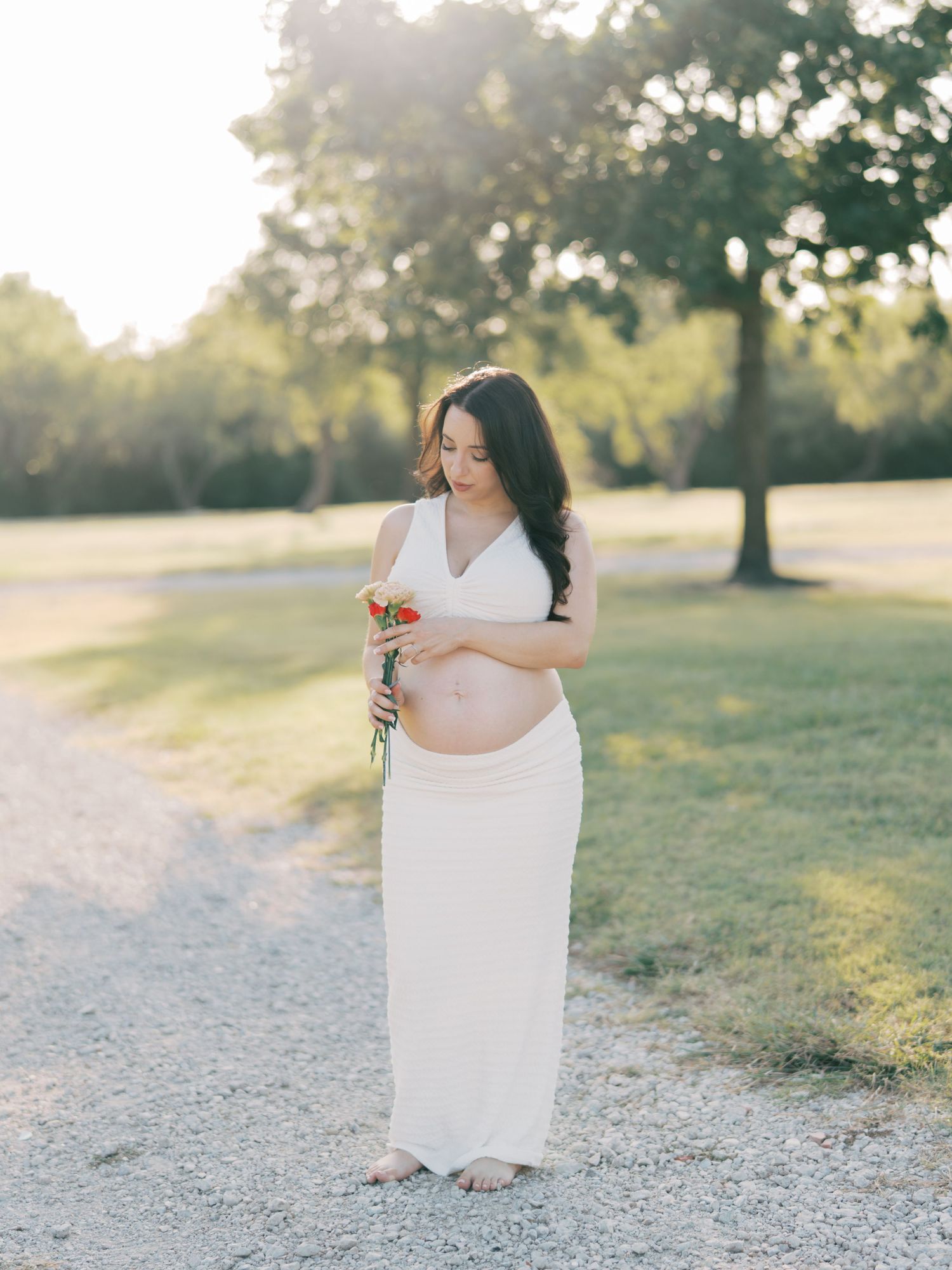 Pregnant woman in white dress holding wildflower bouquet and touching bump in sunny park.