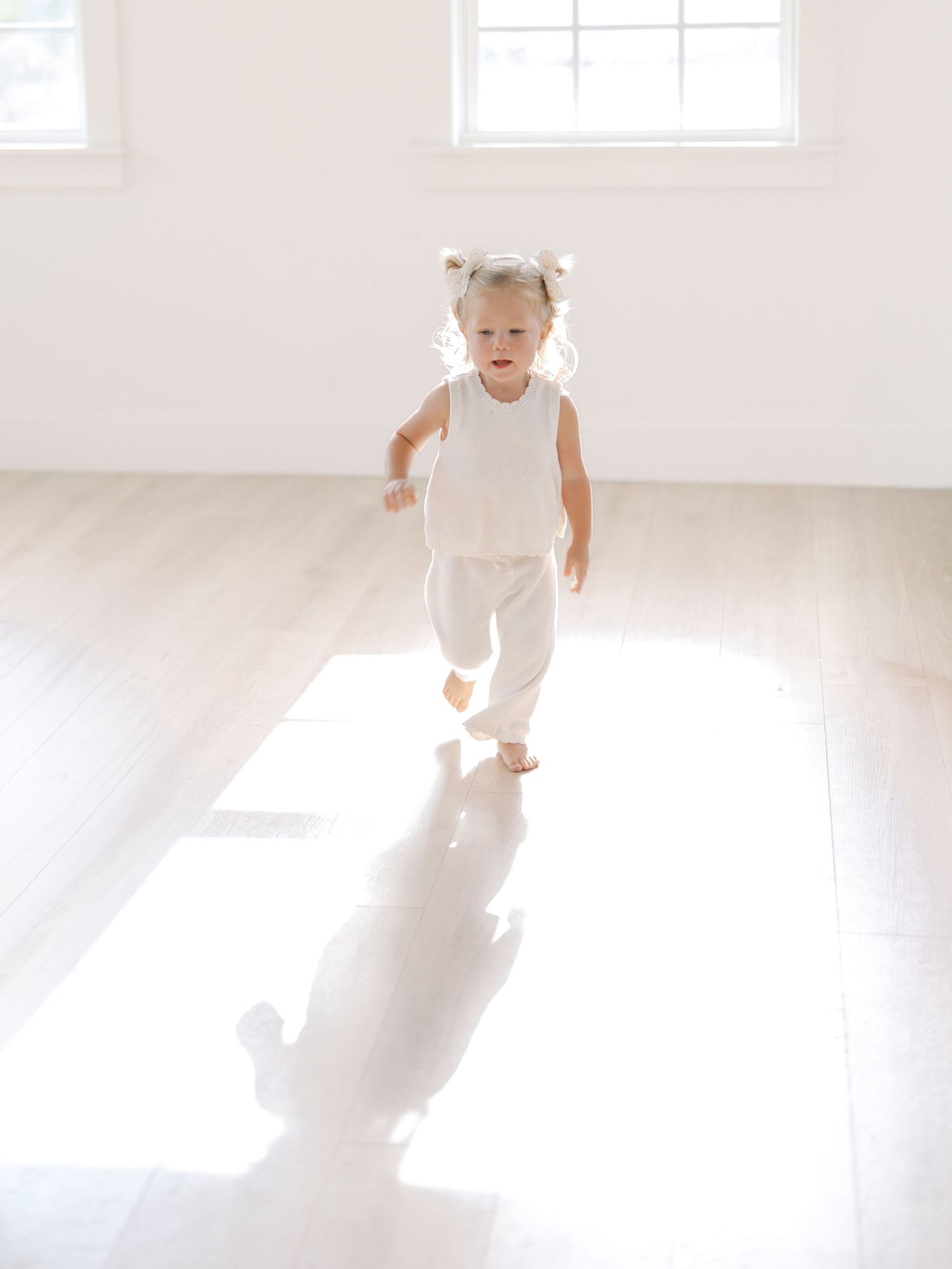 Toddler girl in white outfit running barefoot in a bright white room by Alahir Villasenor Photographer