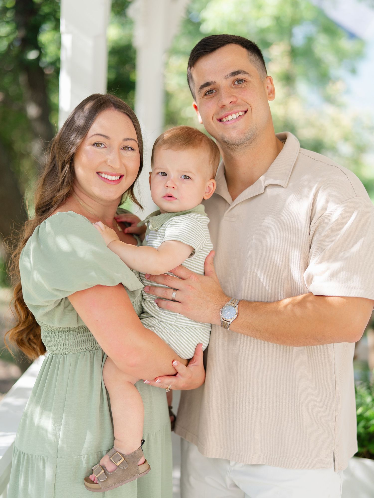 Happy young family with mother in green dress and father in beige shirt holding their baby boy outdoors.