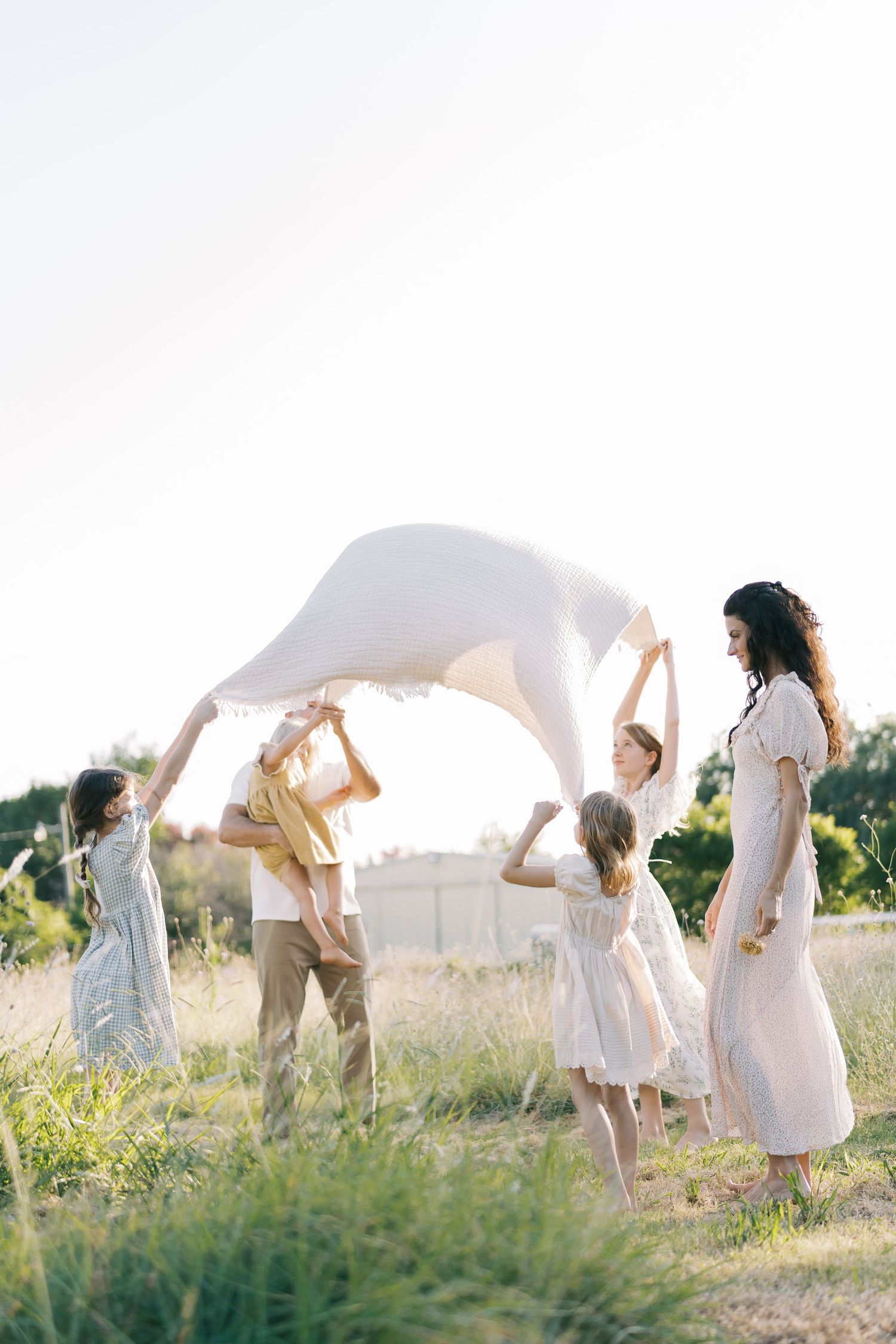 A family joyfully tosses a white blanket in a bright sunlit field, children and parents laughing together.