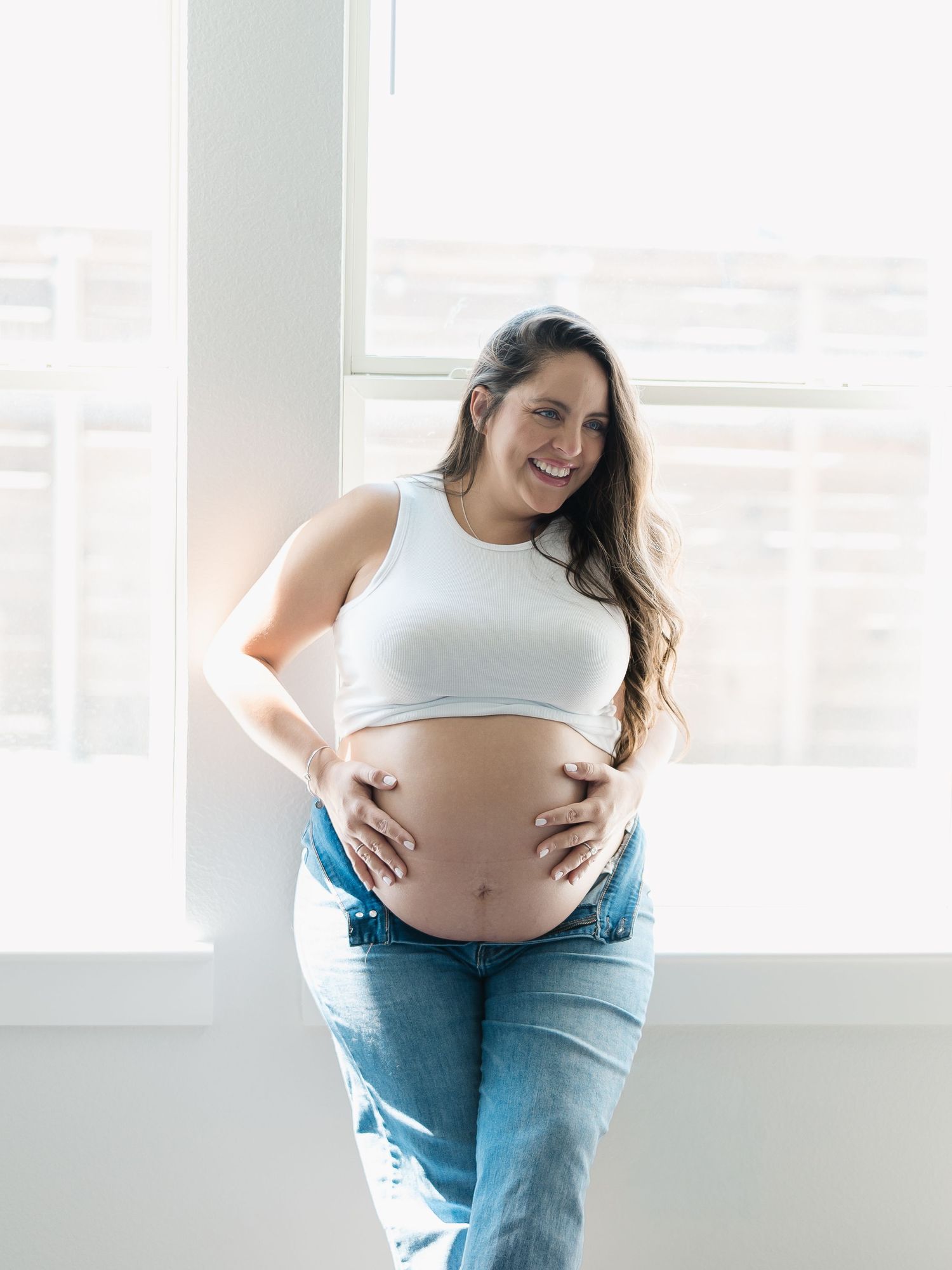 Happy pregnant woman in white crop top and unbuttoned jeans smiling while holding baby bump near bright window by Alahir Villasenor Photogra
