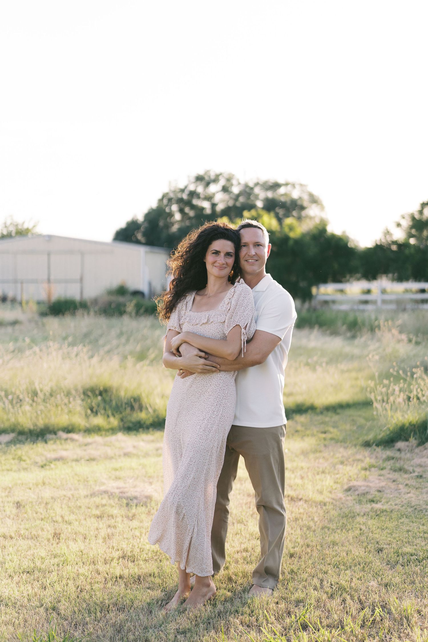 A couple embraces outdoors in a sunlit field, woman in flowy dress, man in white shirt and khaki pants.
