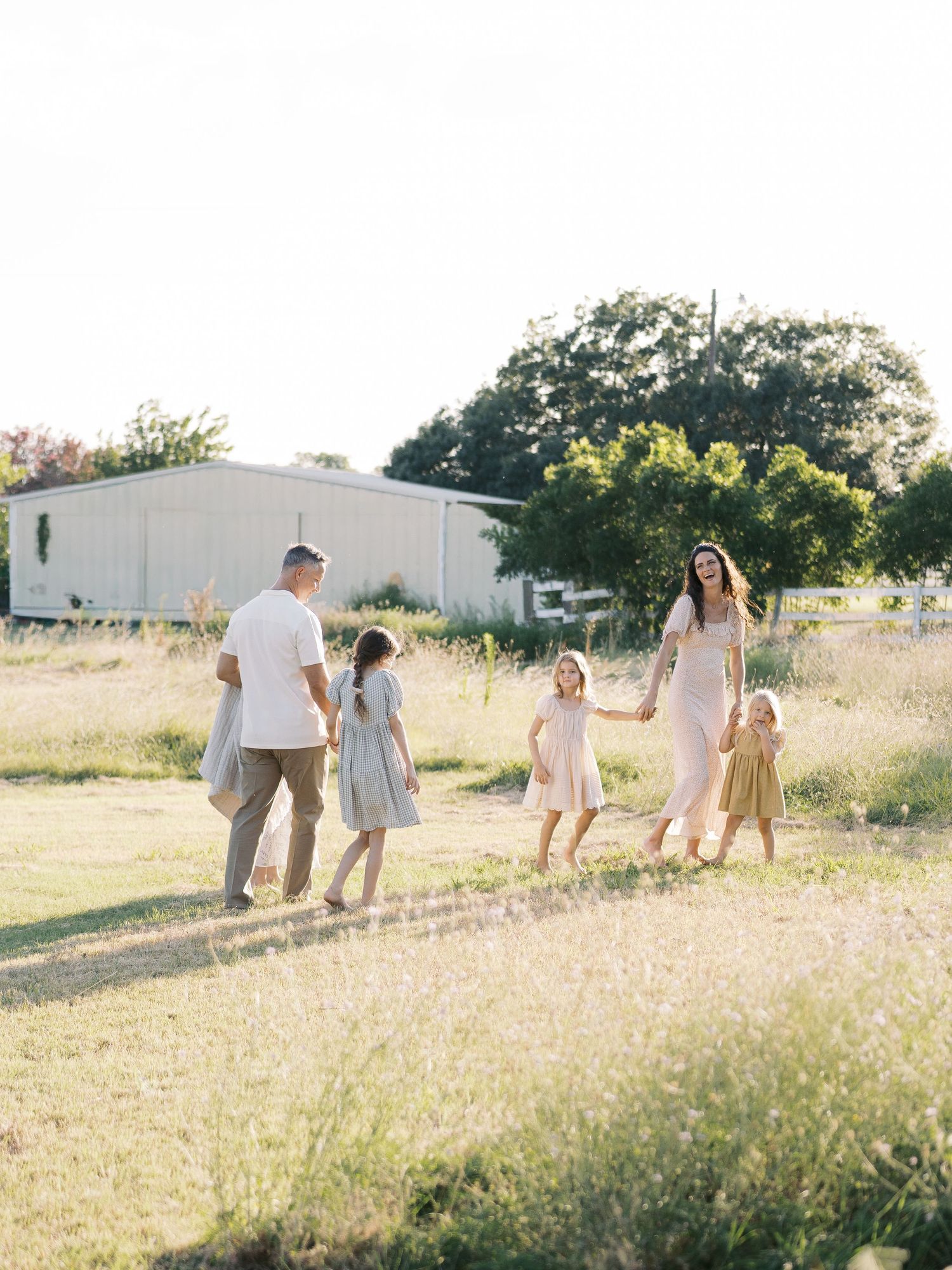 Parents and three daughters holding hands strolling through sunlit grassy field during outdoor family photo session by Alahir Villasenor Pho