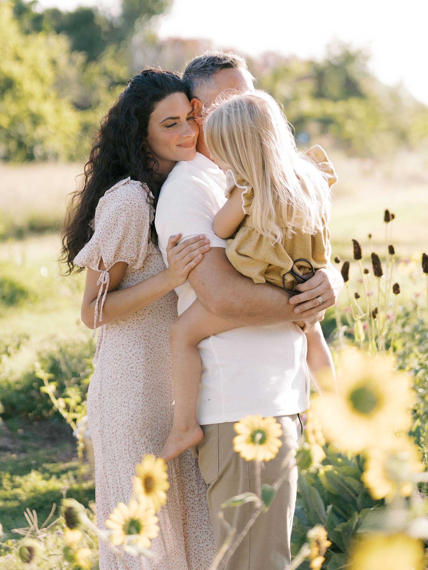 A couple shares a tender moment hugging their young blonde child in a sunflower field at golden hour.