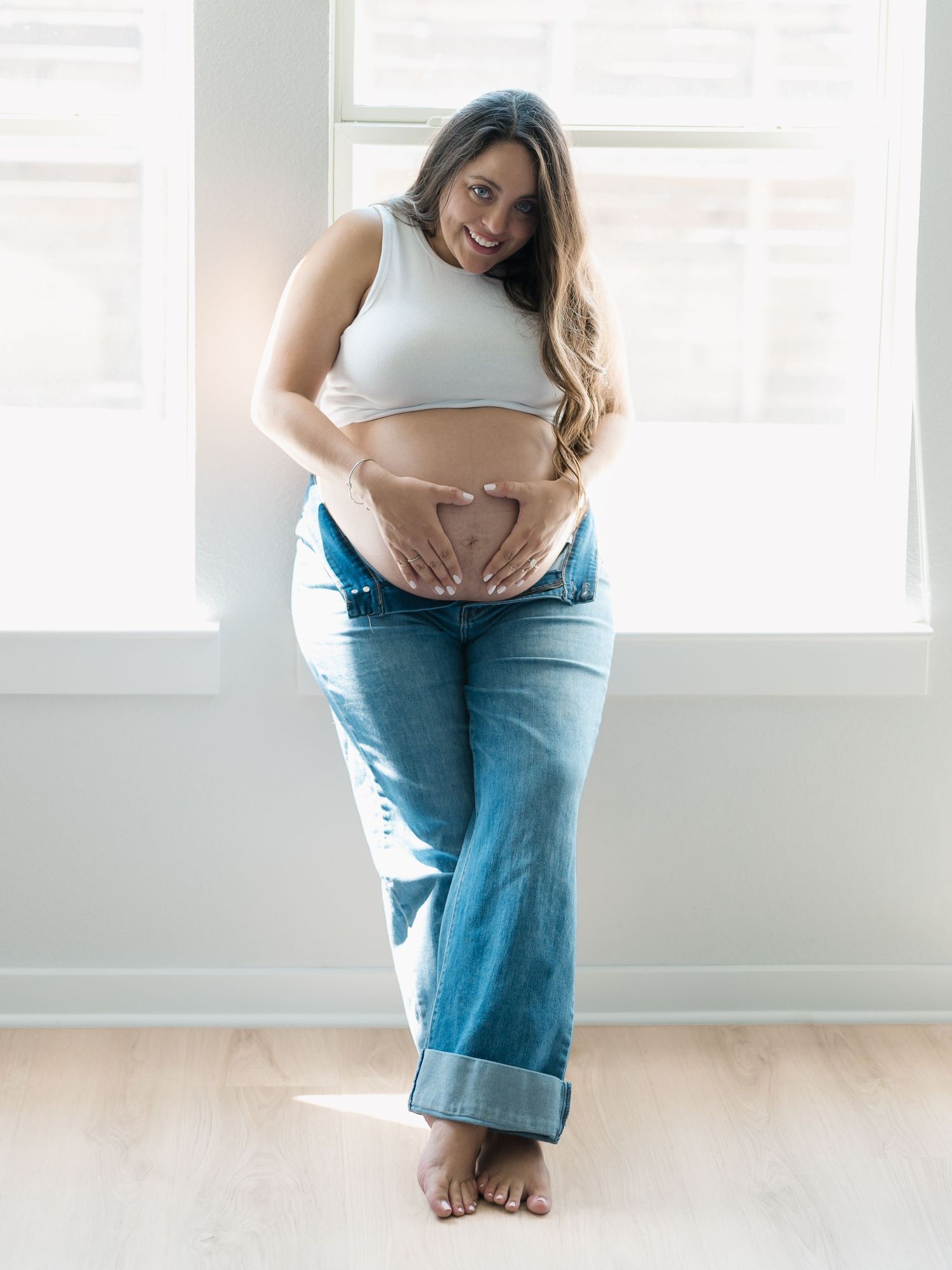 Pregnant woman making heart shape on bare baby bump wearing white crop top and rolled jeans by window by Alahir Villasenor Photographer