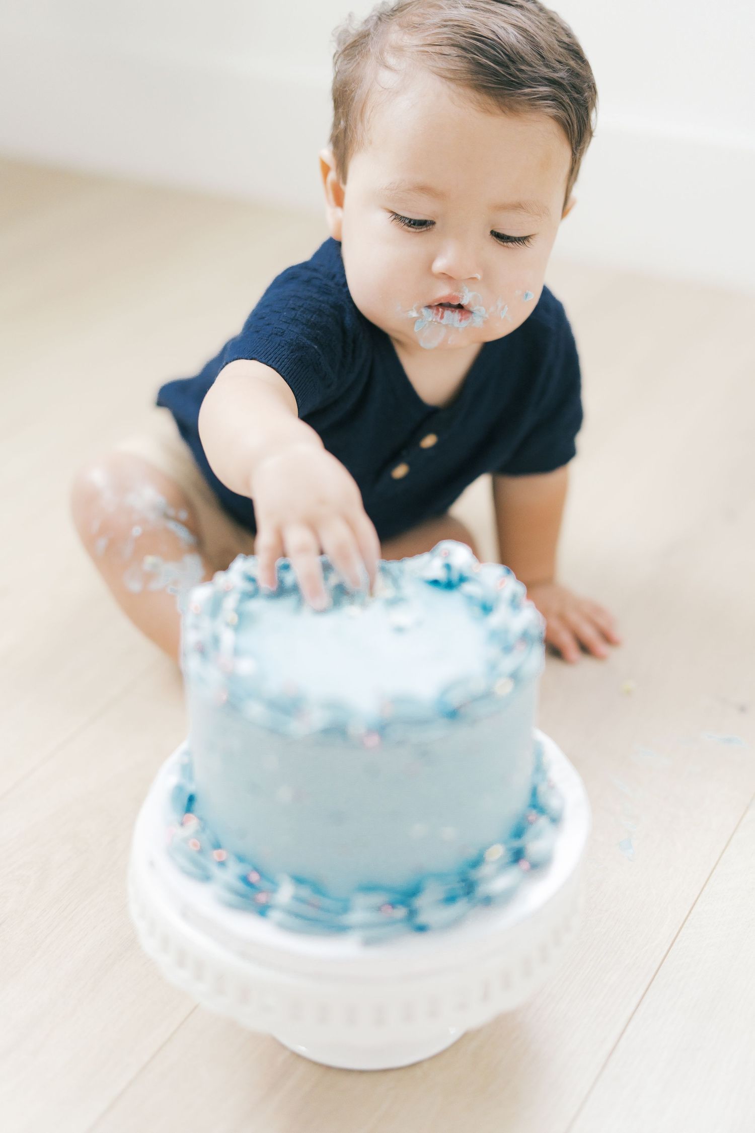 Baby boy in navy onesie smashing a blue confetti cake during first birthday cake smash.