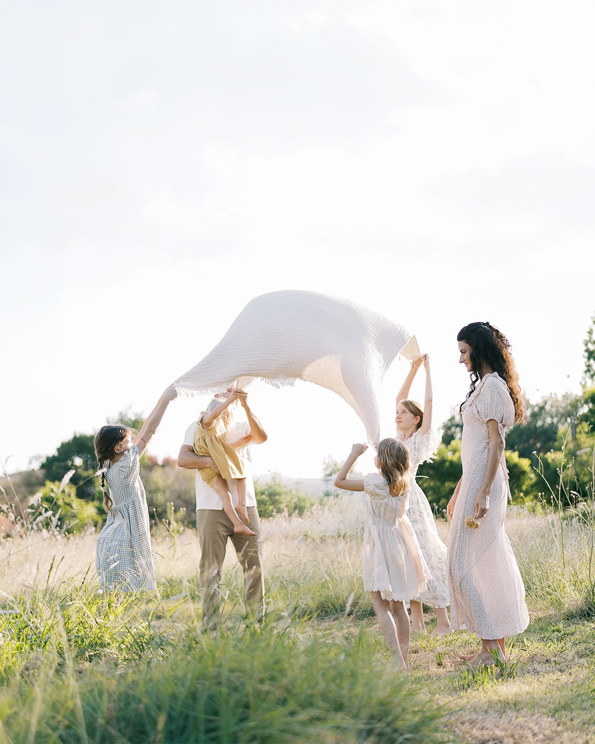 A joyful family tosses a white fabric sheet in a sunlit meadow, children and adults playing together outdoors.