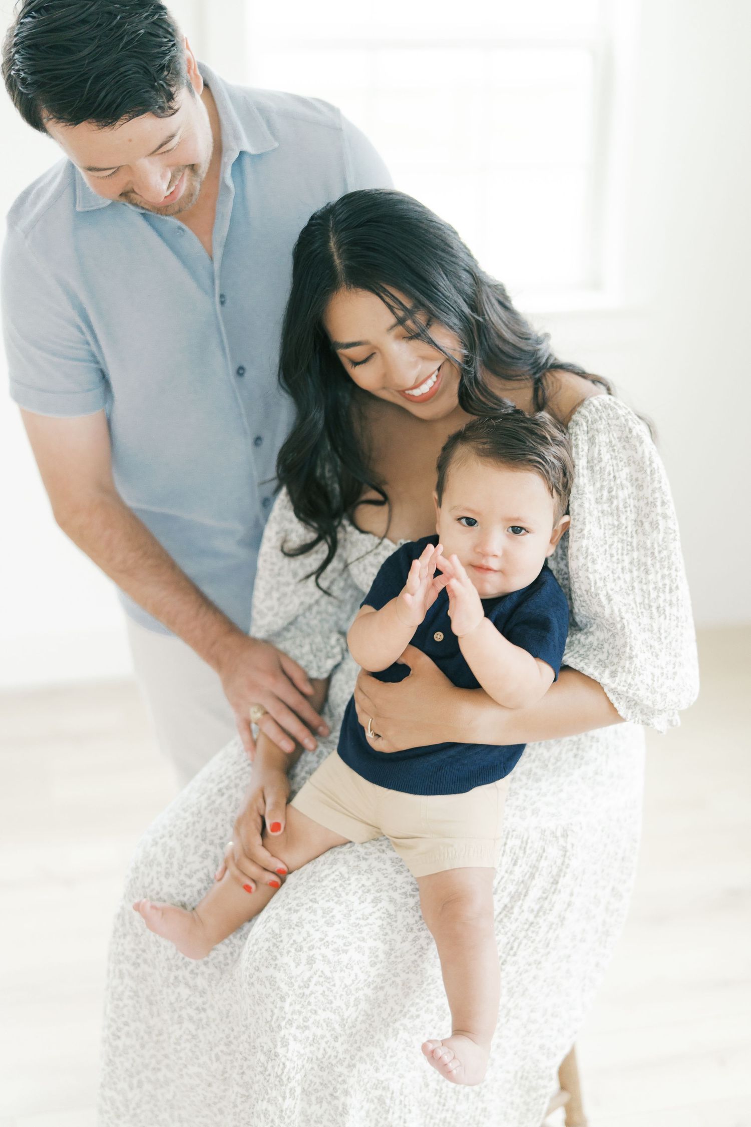 Smiling parents in a bright studio hold their happy baby boy dressed in a navy outfit and khaki shorts.