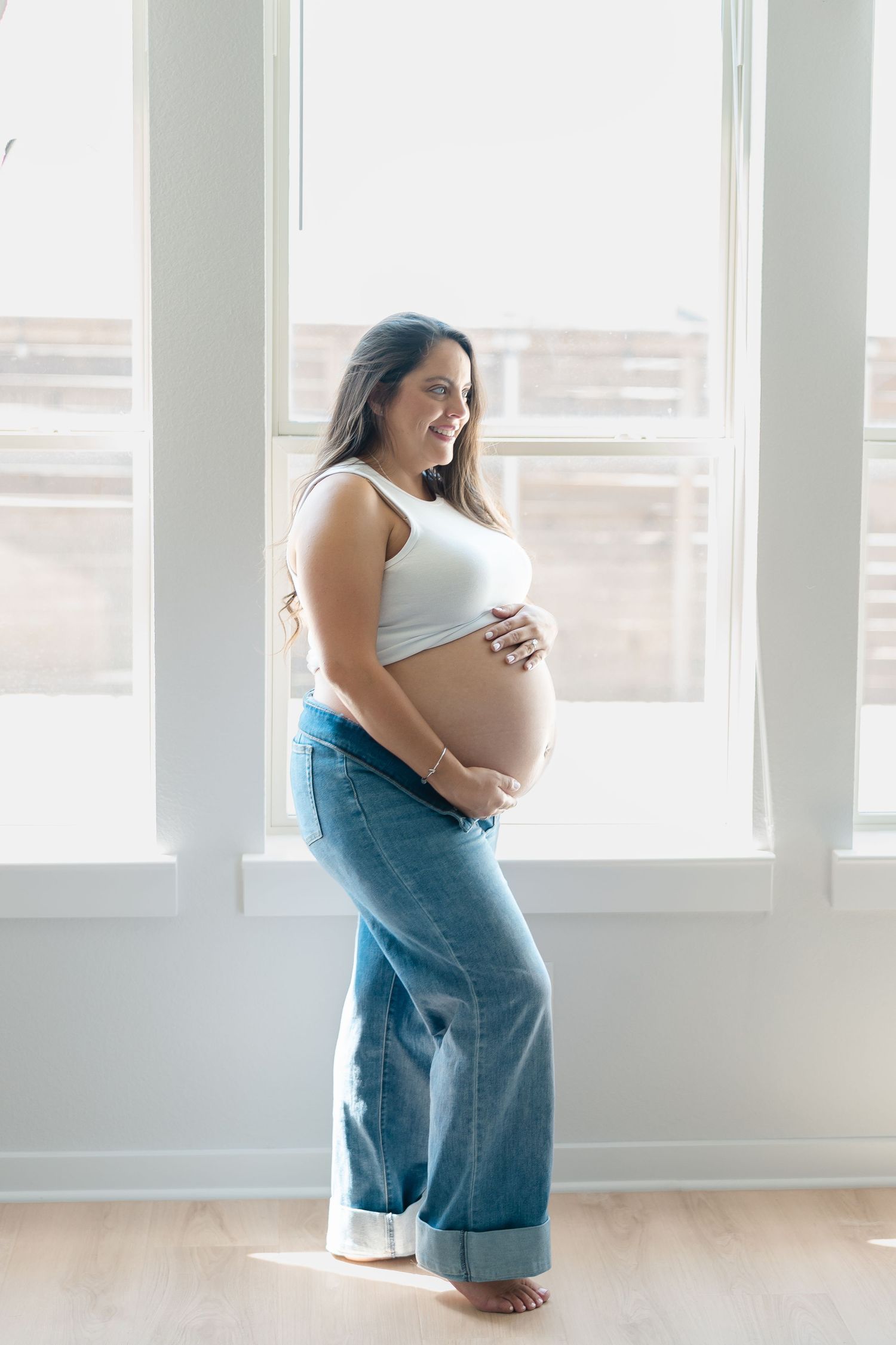 Smiling pregnant woman in white crop top and jeans cradling bare baby bump by bright window indoors.