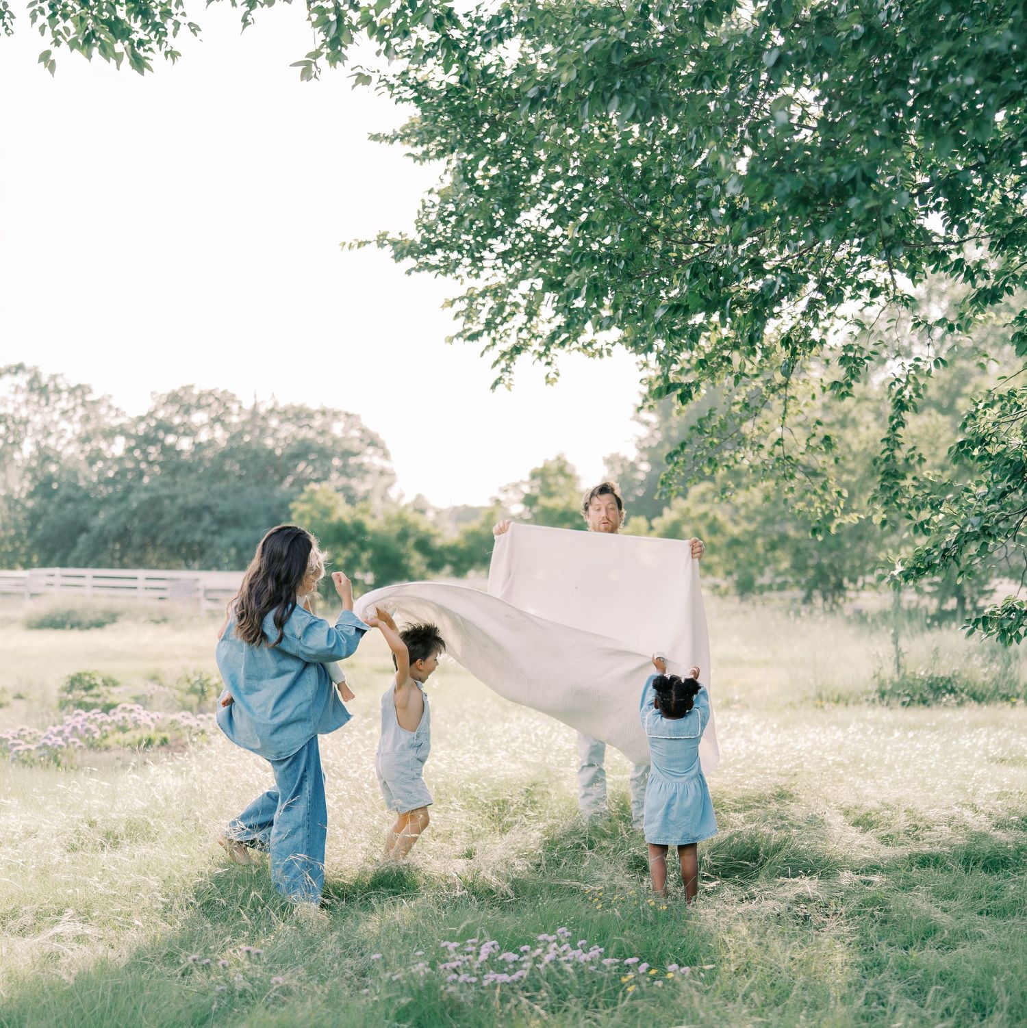 A family spreads a white blanket in a sunlit meadow beneath a large green tree in Dallas by Alahir Villasenor Photography.