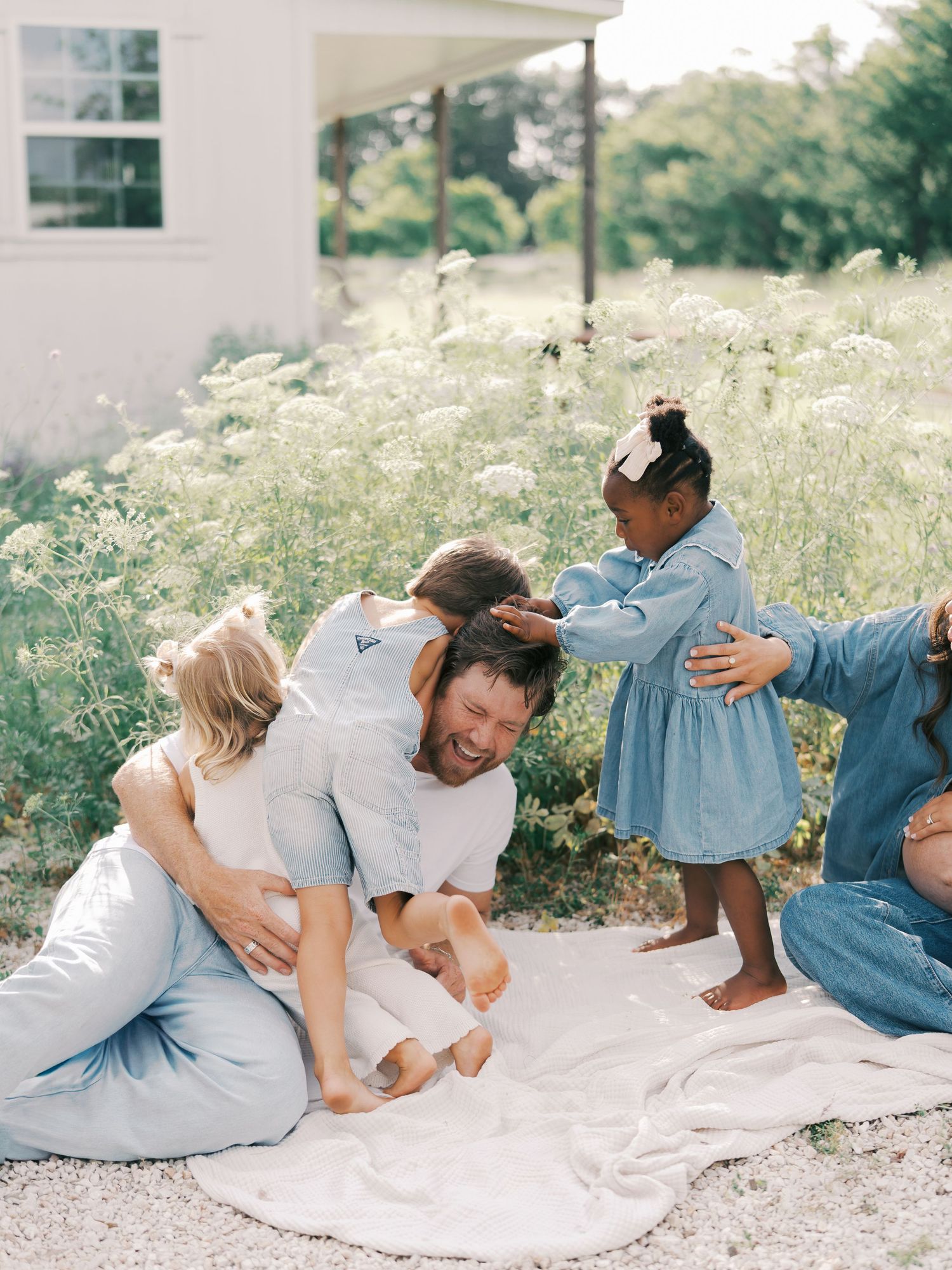 A joyful family plays together on a blanket outdoors, with children climbing on a laughing father near wildflowers.