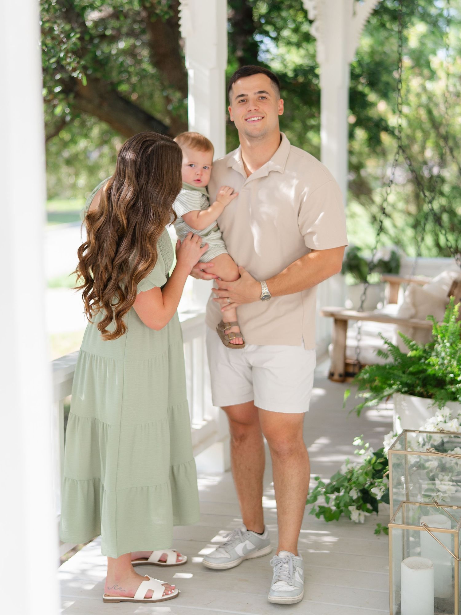 A young family poses on a white porch, mother in sage dress holding a baby, father in beige polo and white shorts.