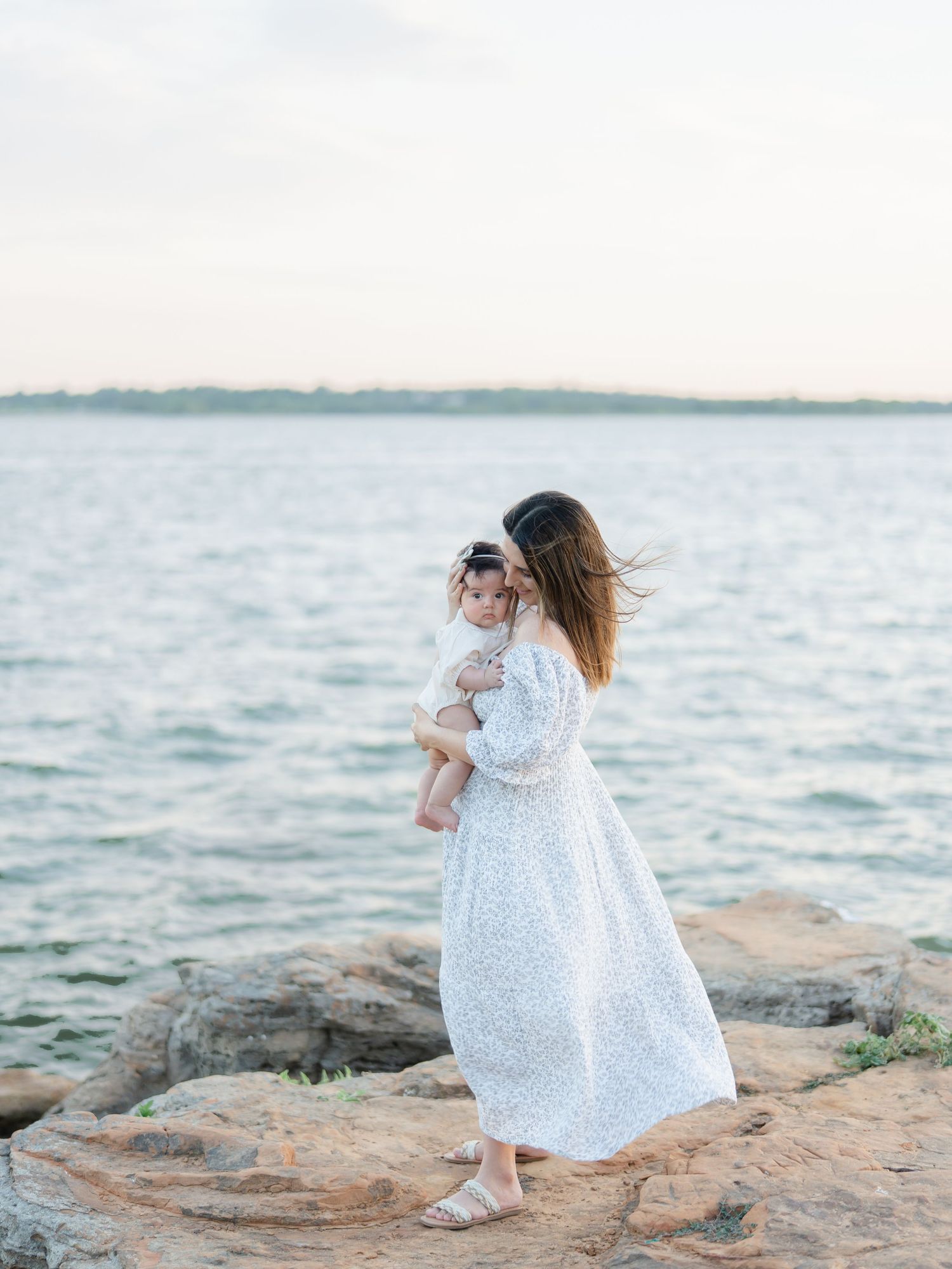 A mother in a white floral dress holds her baby on rocky shores by calm water at sunset.