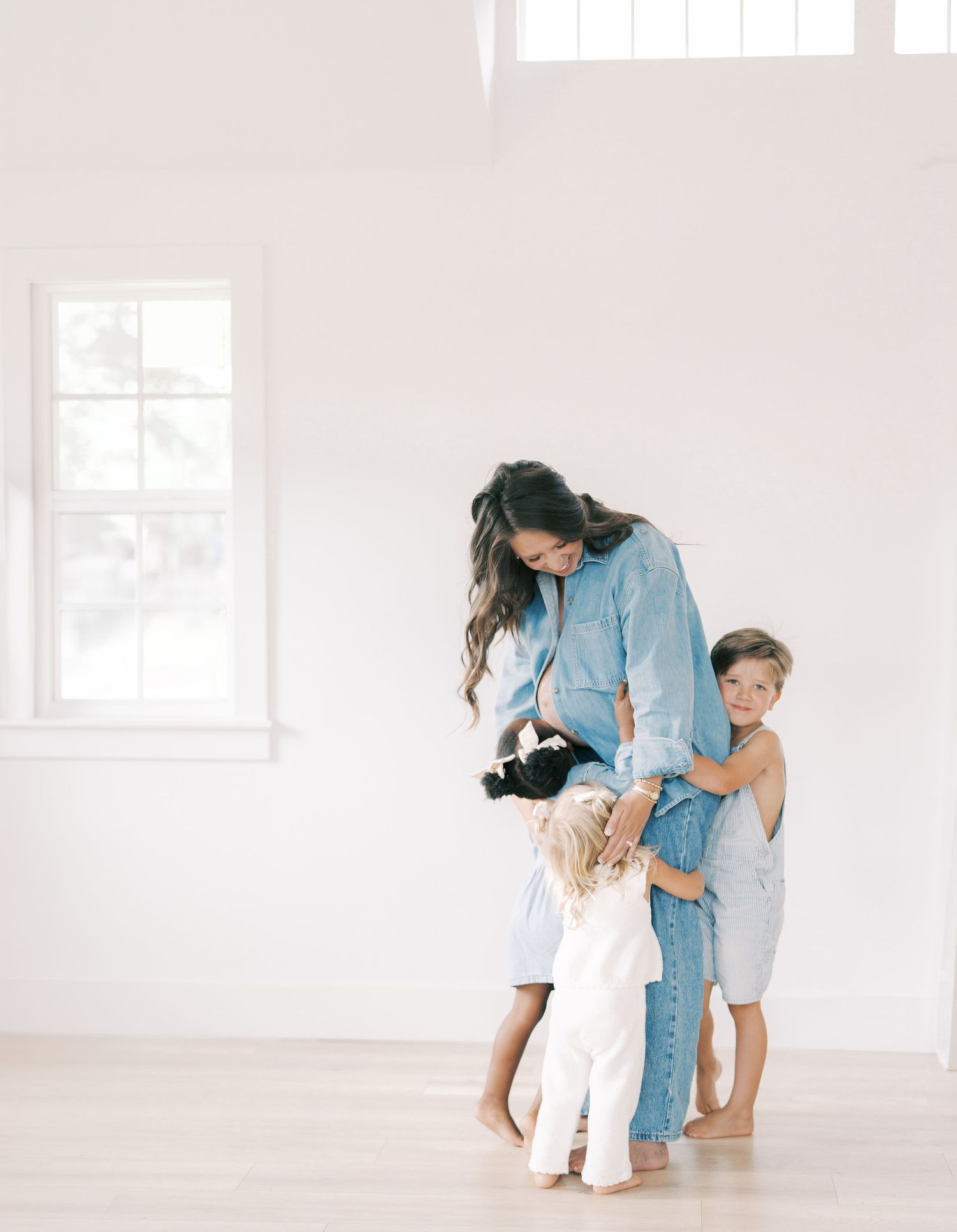 A mother in blue denim embraces her three children in a bright white minimalist room in Dallas by Alahir Villasenor Photography.
