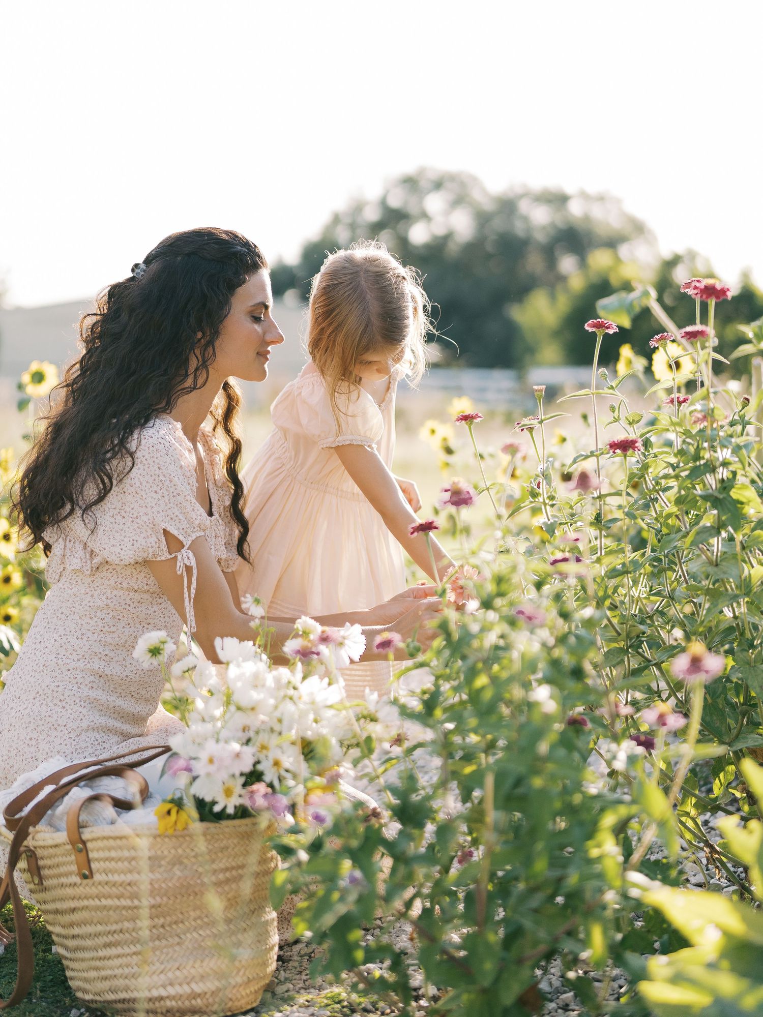 Mother and young daughter in floral dresses picking flowers in a sunlit garden at golden hour.