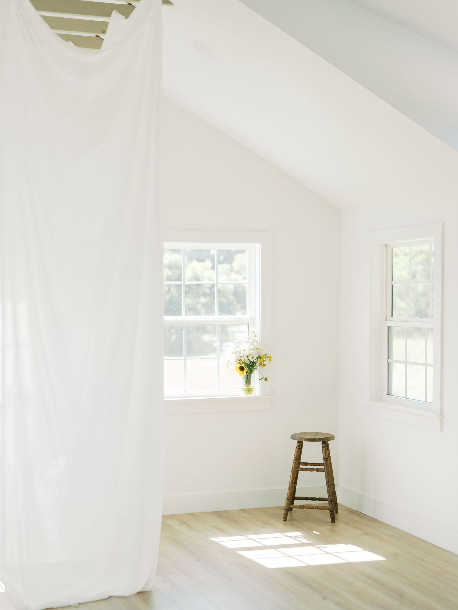 A bright white room with vaulted ceilings, sheer curtains, and a wooden stool by windows in Dallas by Alahir Villasenor Photography.