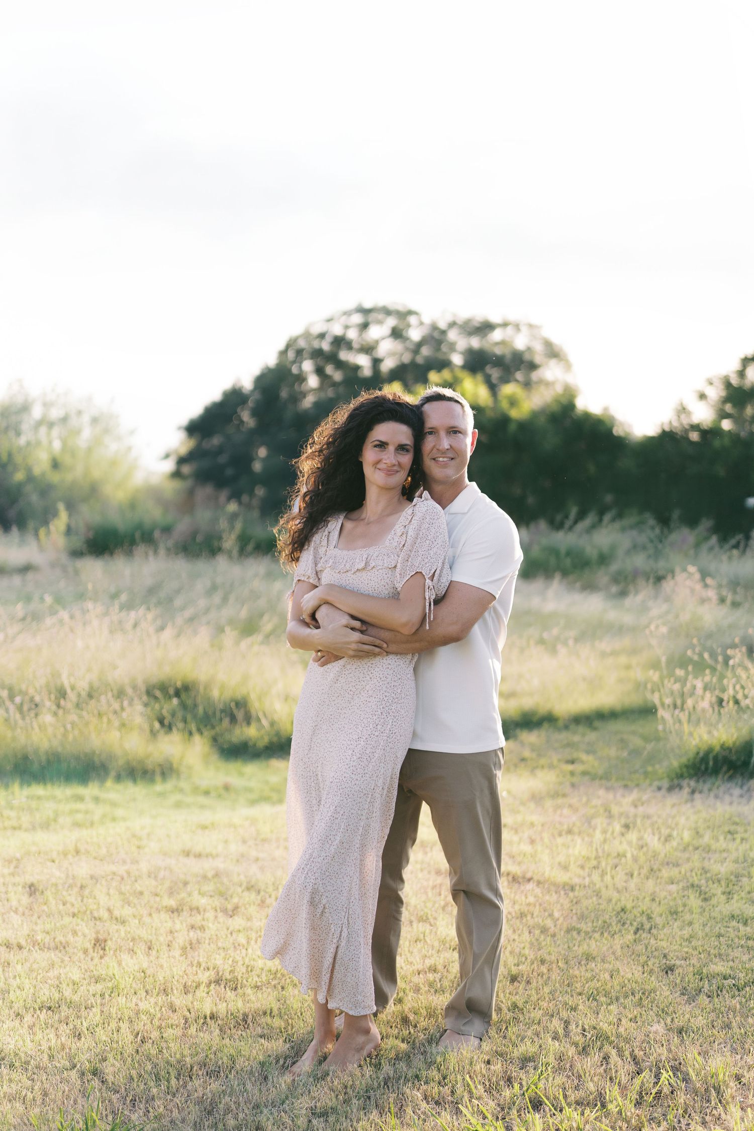 A couple embraces in a sunlit field, the woman in a flowy beige dress and the man in a white shirt and khaki pants.