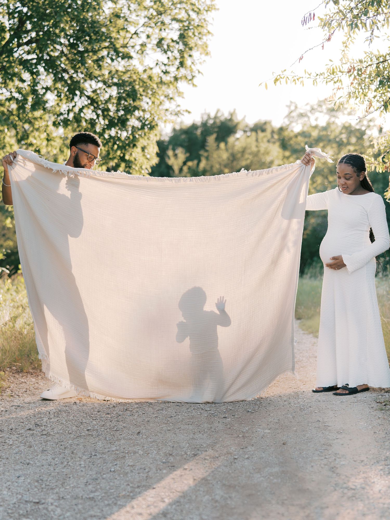 Couple holding white fabric sheet revealing child silhouette shadow during creative maternity photo shoot outdoors.