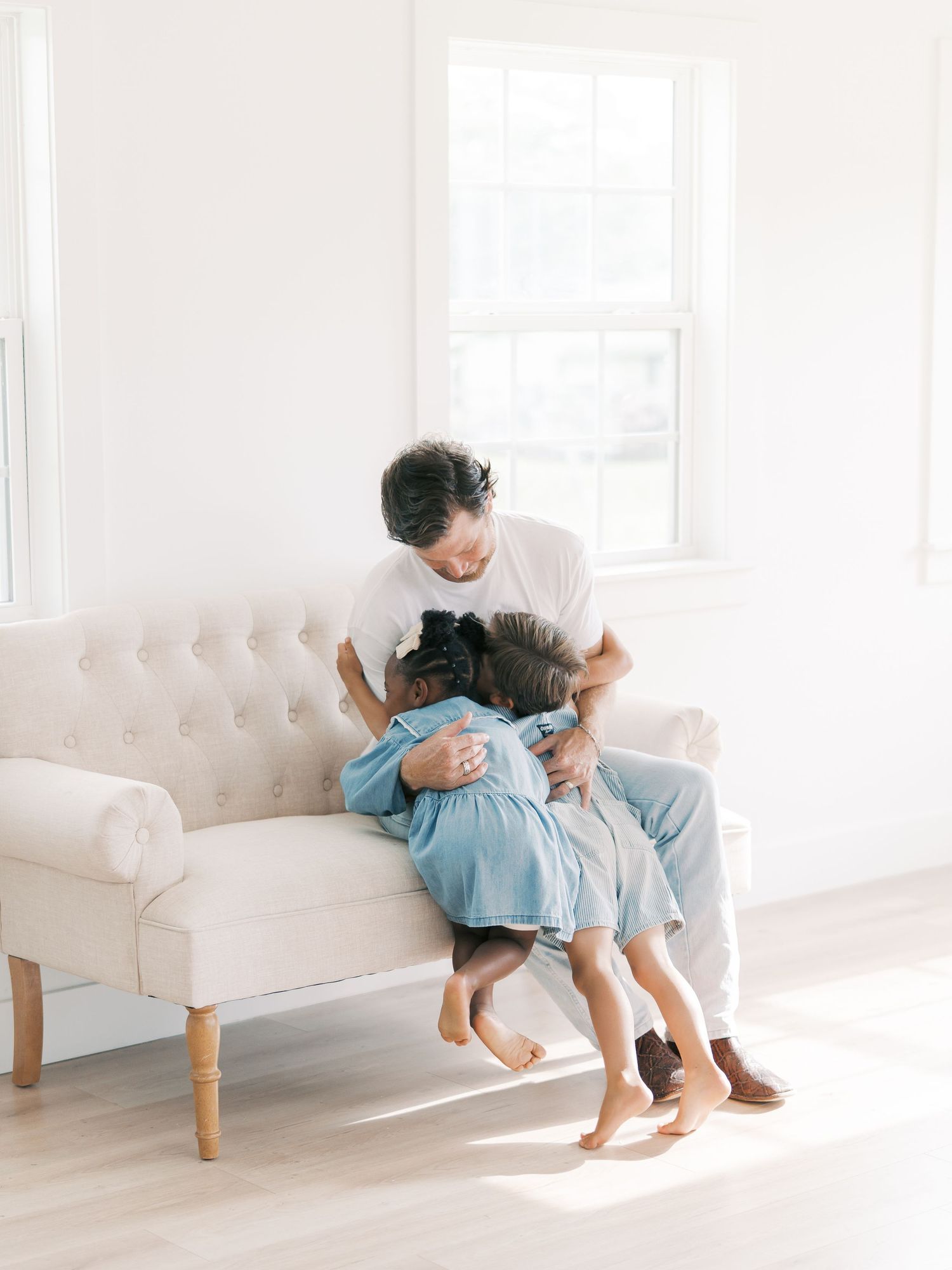 A father tenderly embraces his young daughter on a white sofa in a sunlit room by Alahir Villasenor Photographer