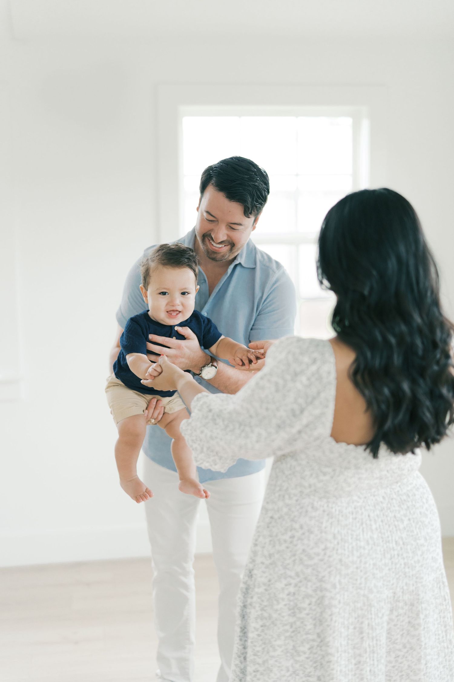 A smiling father in a light blue shirt holds his baby boy while the mother reaches out toward them indoors.