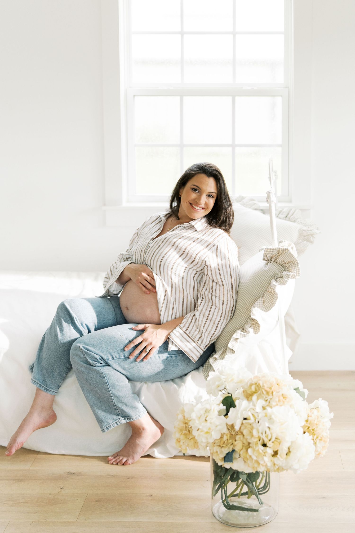 Pregnant woman in striped blouse sitting on white daybed with hydrangeas in bright studio.
