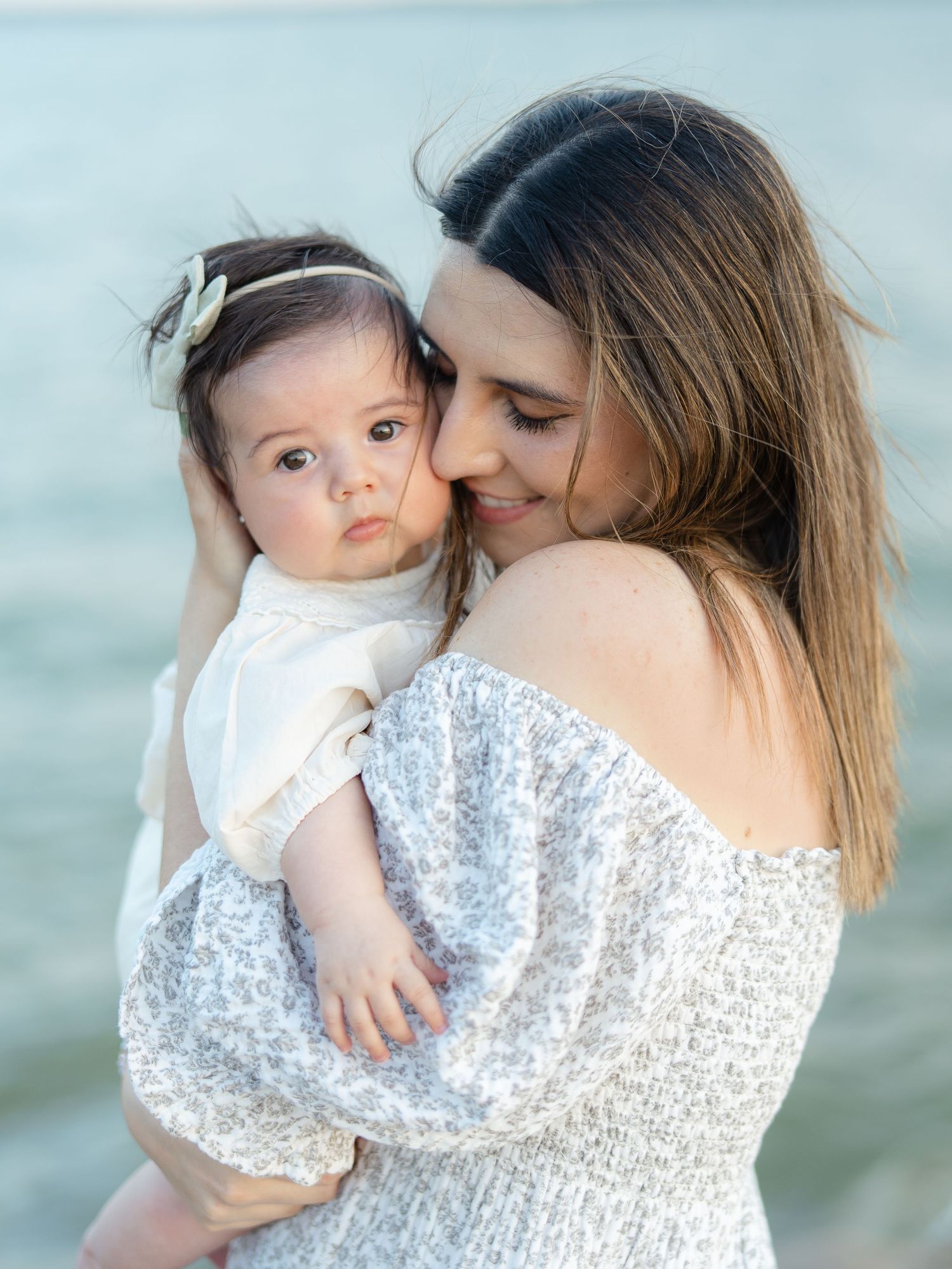 A mother tenderly holds her baby girl near the water, both wearing white in a serene outdoor portrait.