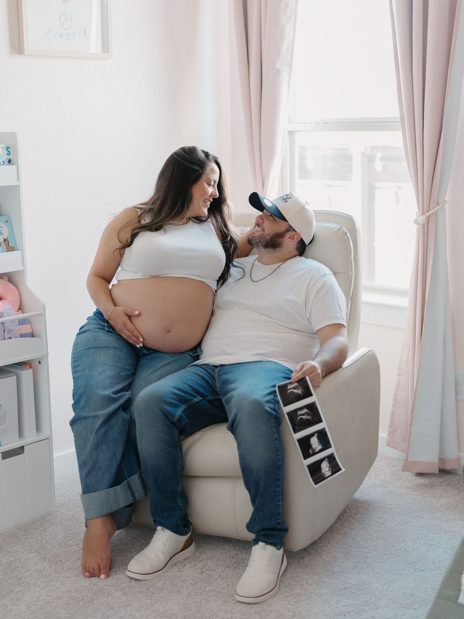 Pregnant couple in nursery, woman standing with bare bump as man sits in recliner holding ultrasound photos. by Alahir Villasenor Photograph