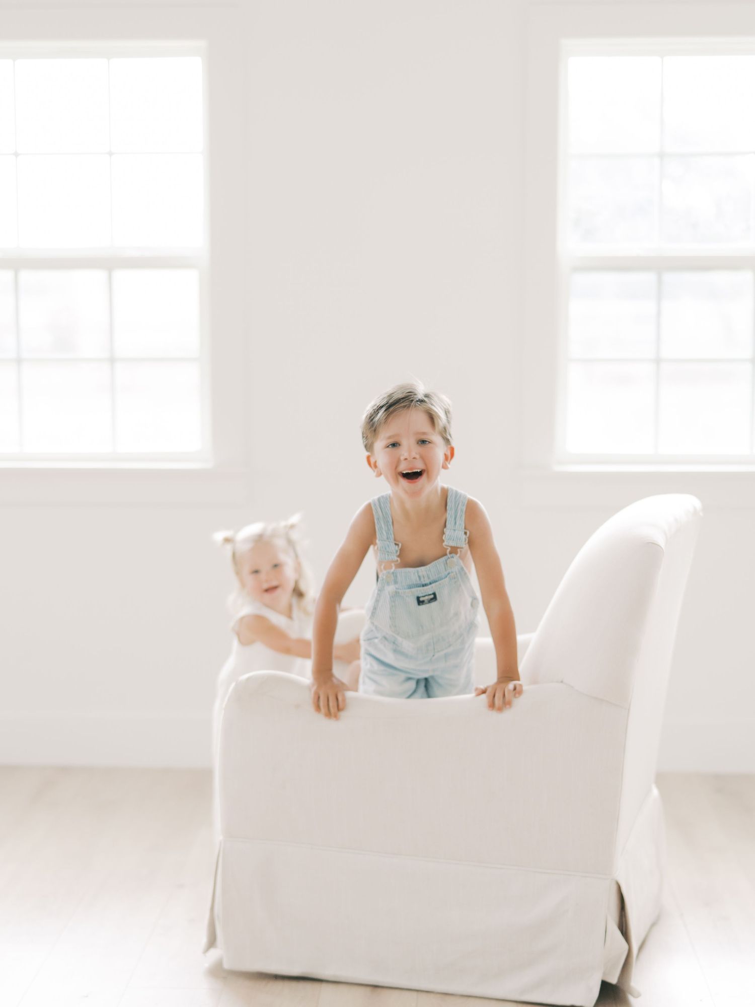 Two young children laugh and play on a white armchair in a bright studio setting by Alahir Villasenor Photographer
