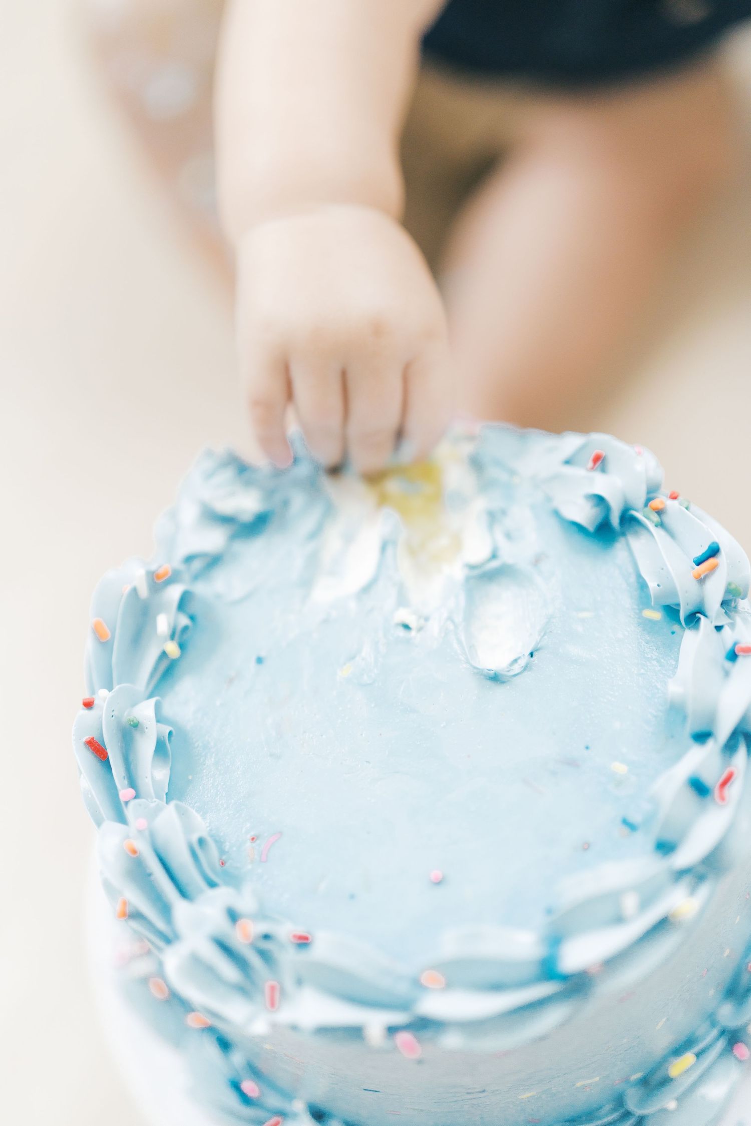 A baby's small hand reaches into a blue frosted smash cake decorated with colorful sprinkles for a first birthday.