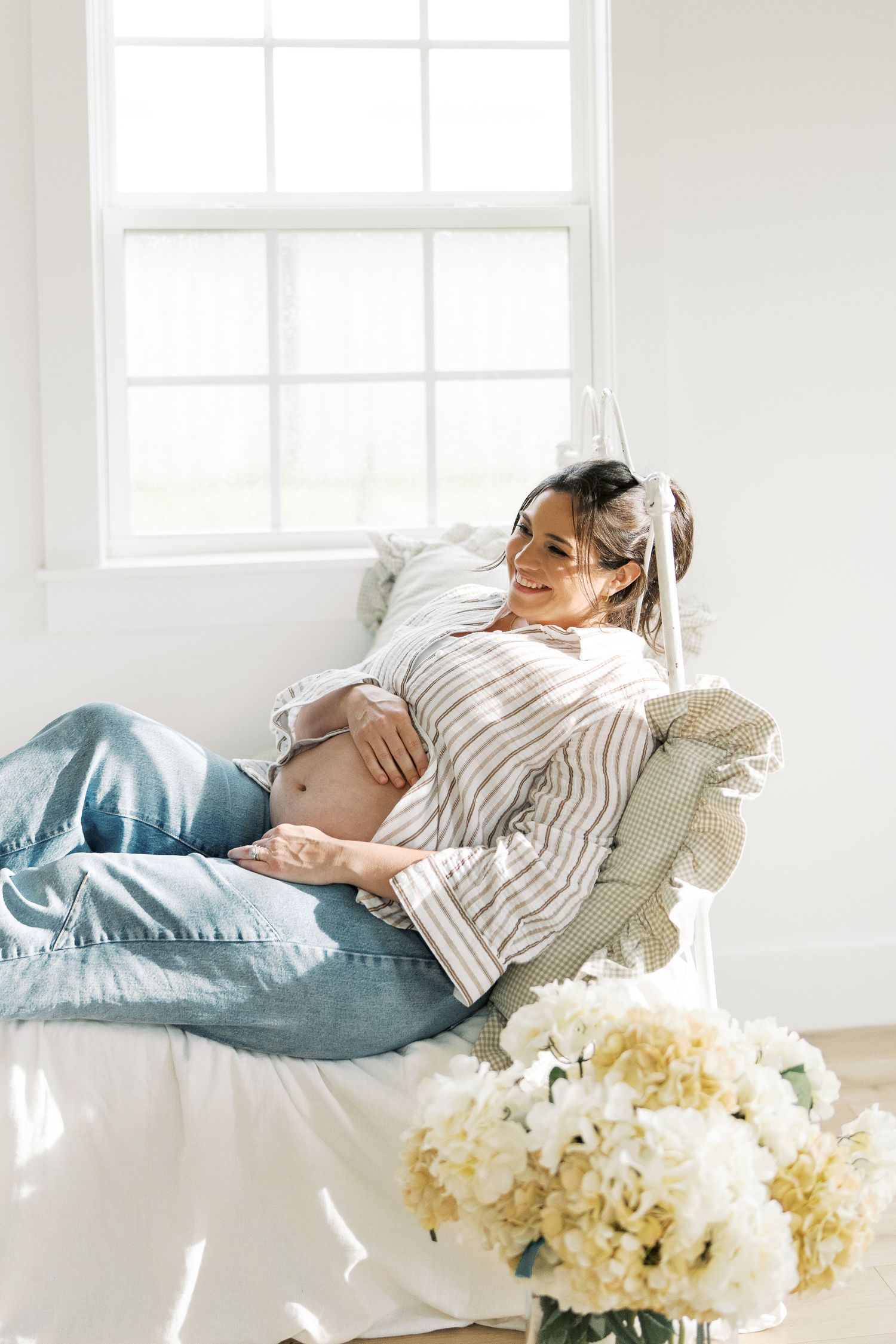 Pregnant woman in striped blouse and jeans relaxing on white daybed with hydrangeas nearby.