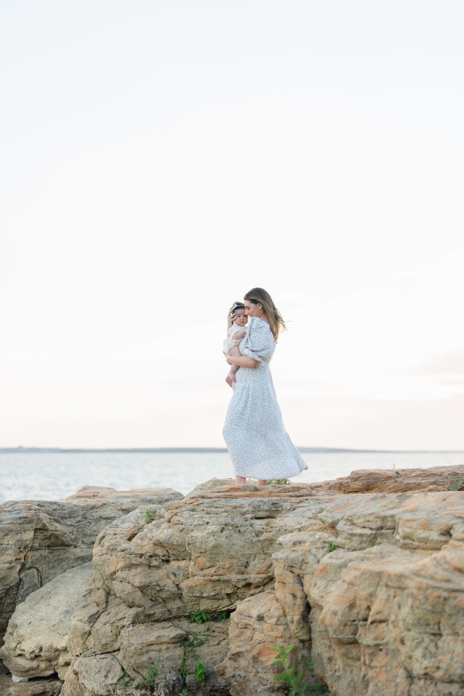 Mother in white maxi dress holding baby on rocky shoreline with calm ocean in background.