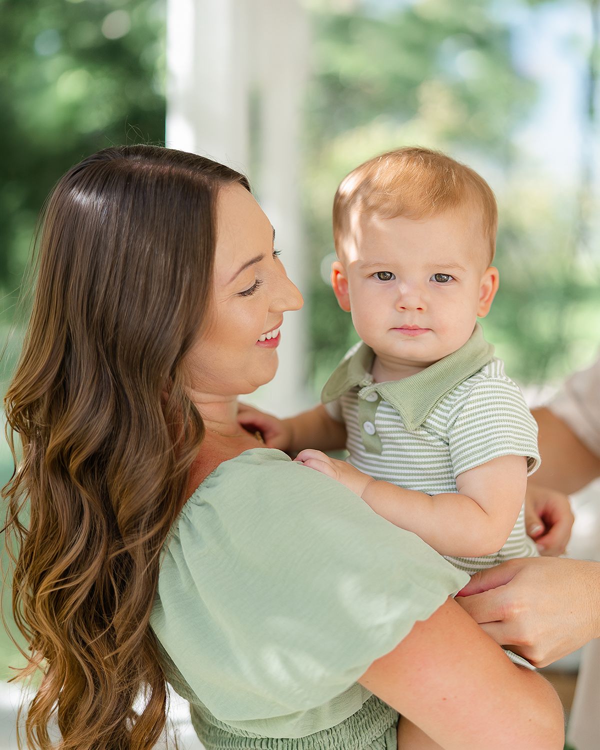 A smiling woman with long brown hair holds a cute baby boy in a green striped polo shirt near a bright window.