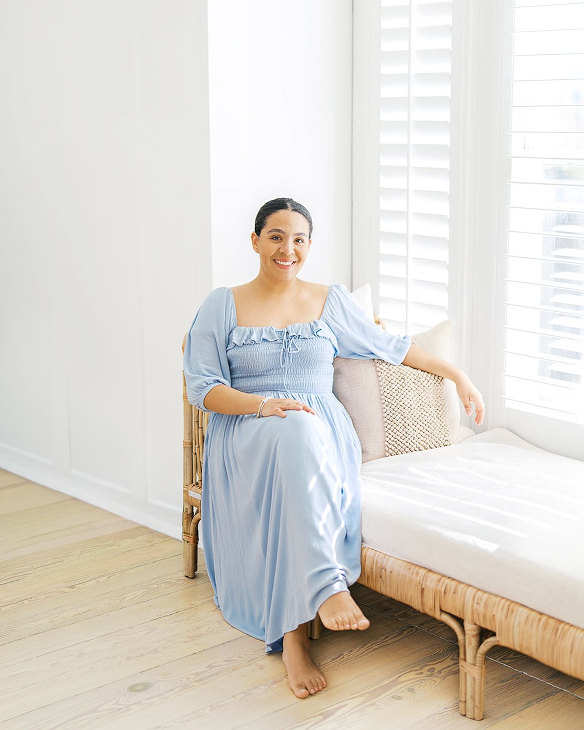 Pregnant woman in light blue ruffle dress sitting on rattan daybed near white shuttered window on light wood floor.