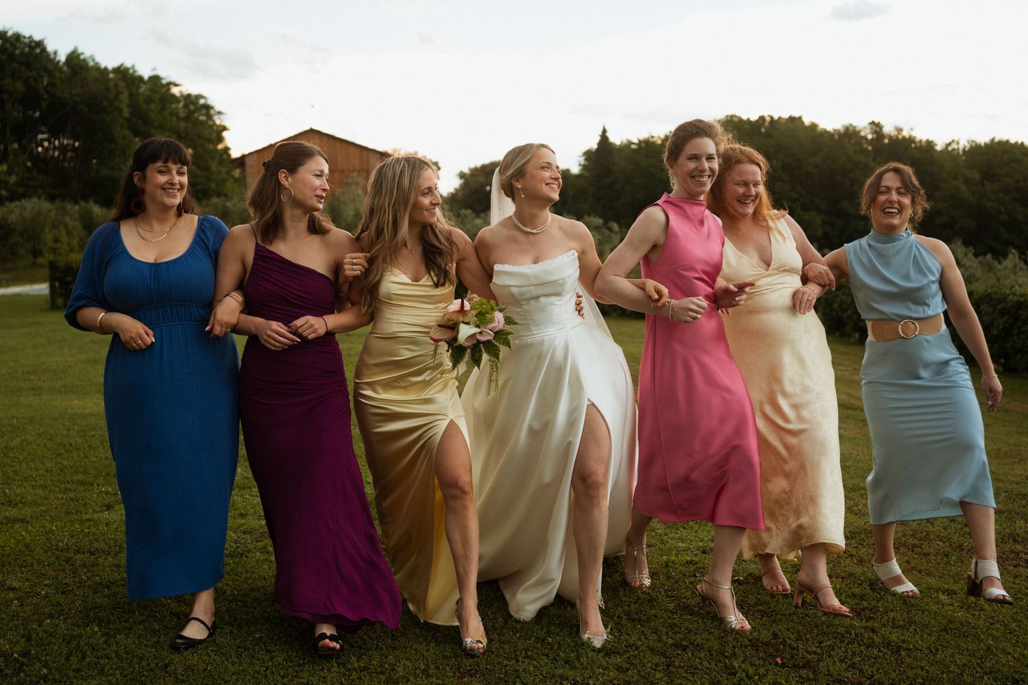 Bride and six bridesmaids in colorful mismatched gowns walking and laughing together outdoors.
