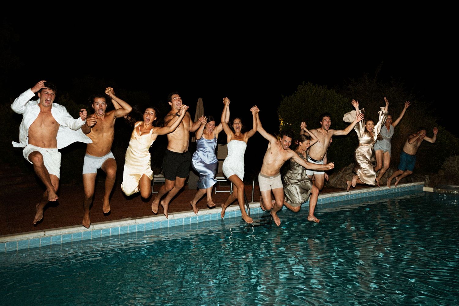 Wedding guests jumping into pool together at night in joyful celebration photo.