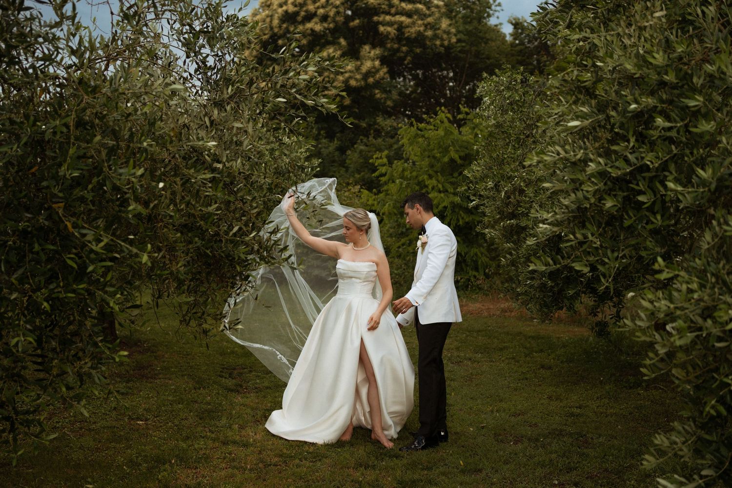 A bride and groom dance together among lush green trees as her veil billows in the wind.