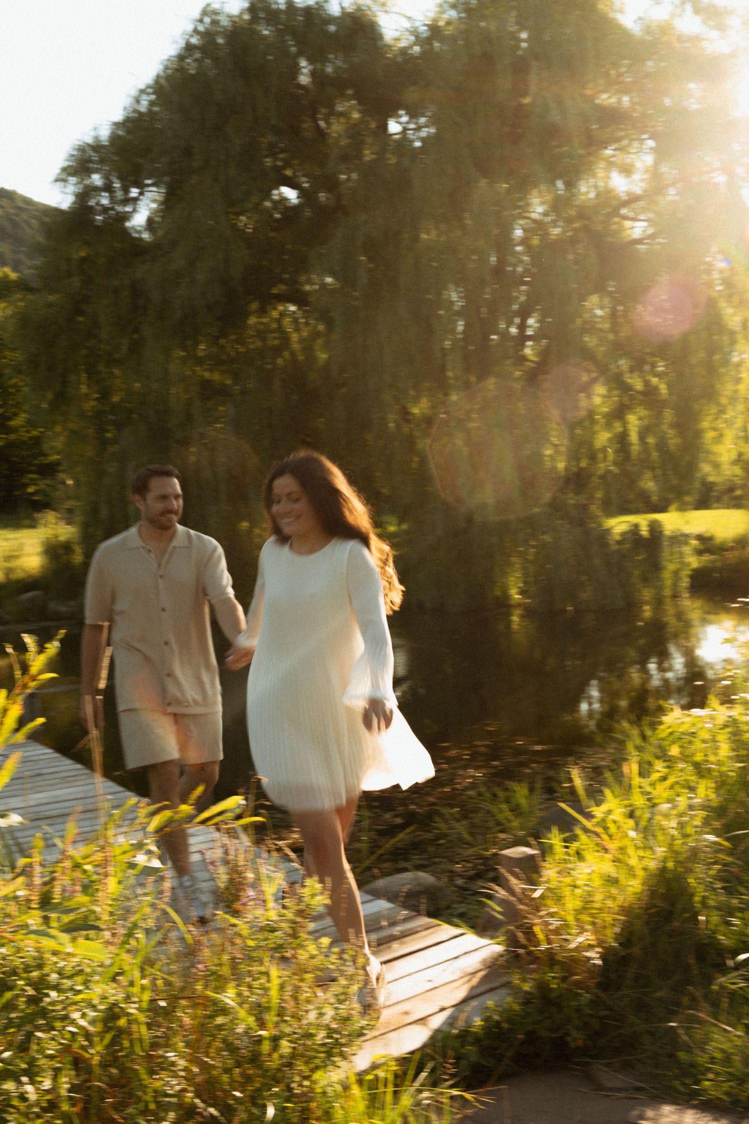 Couple in white outfits walking hand-in-hand on a wooden dock by a sunlit pond surrounded by lush greenery.