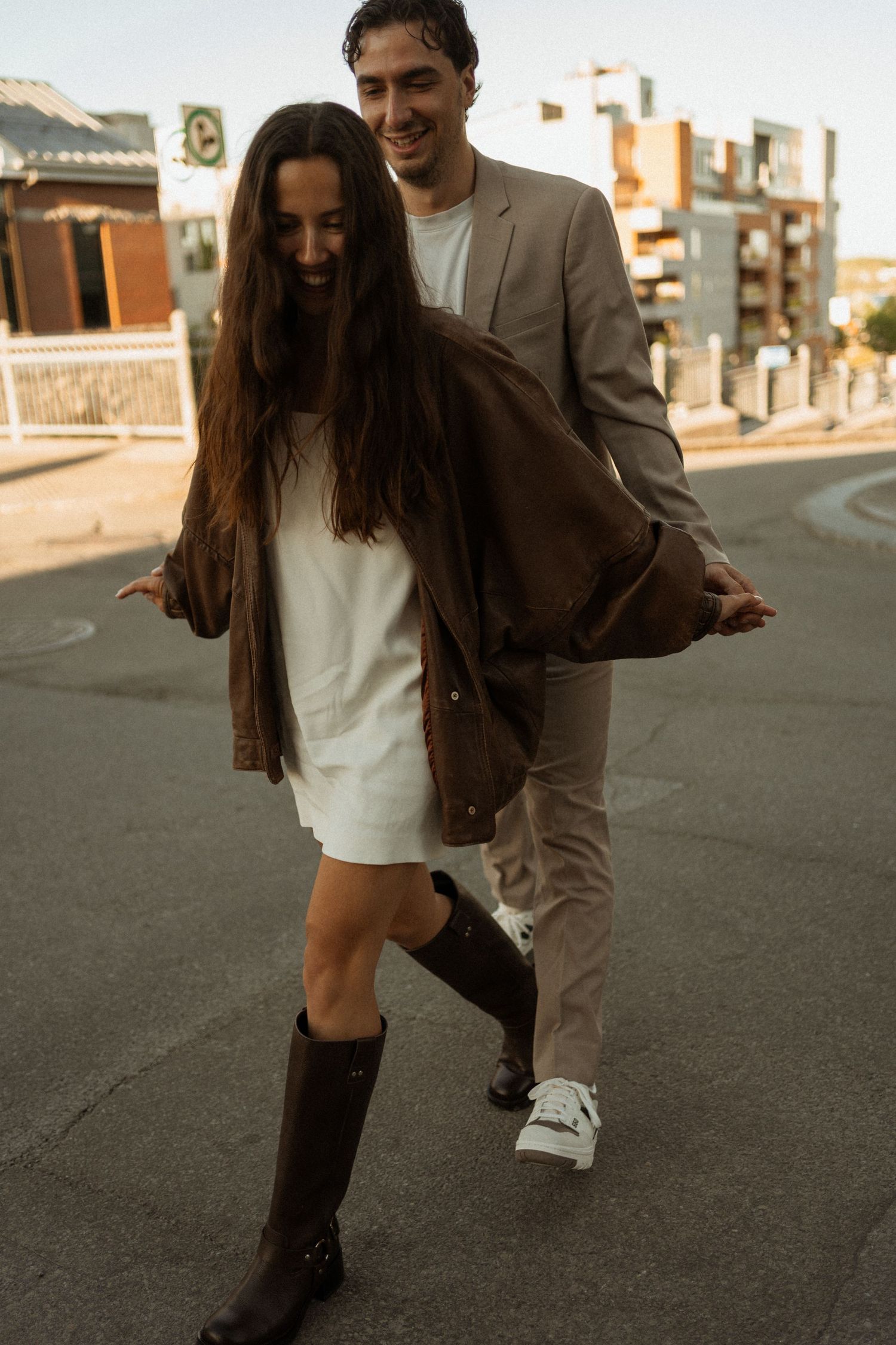 A smiling couple walks on a street, woman in white dress, brown jacket and knee-high boots, man in beige suit.