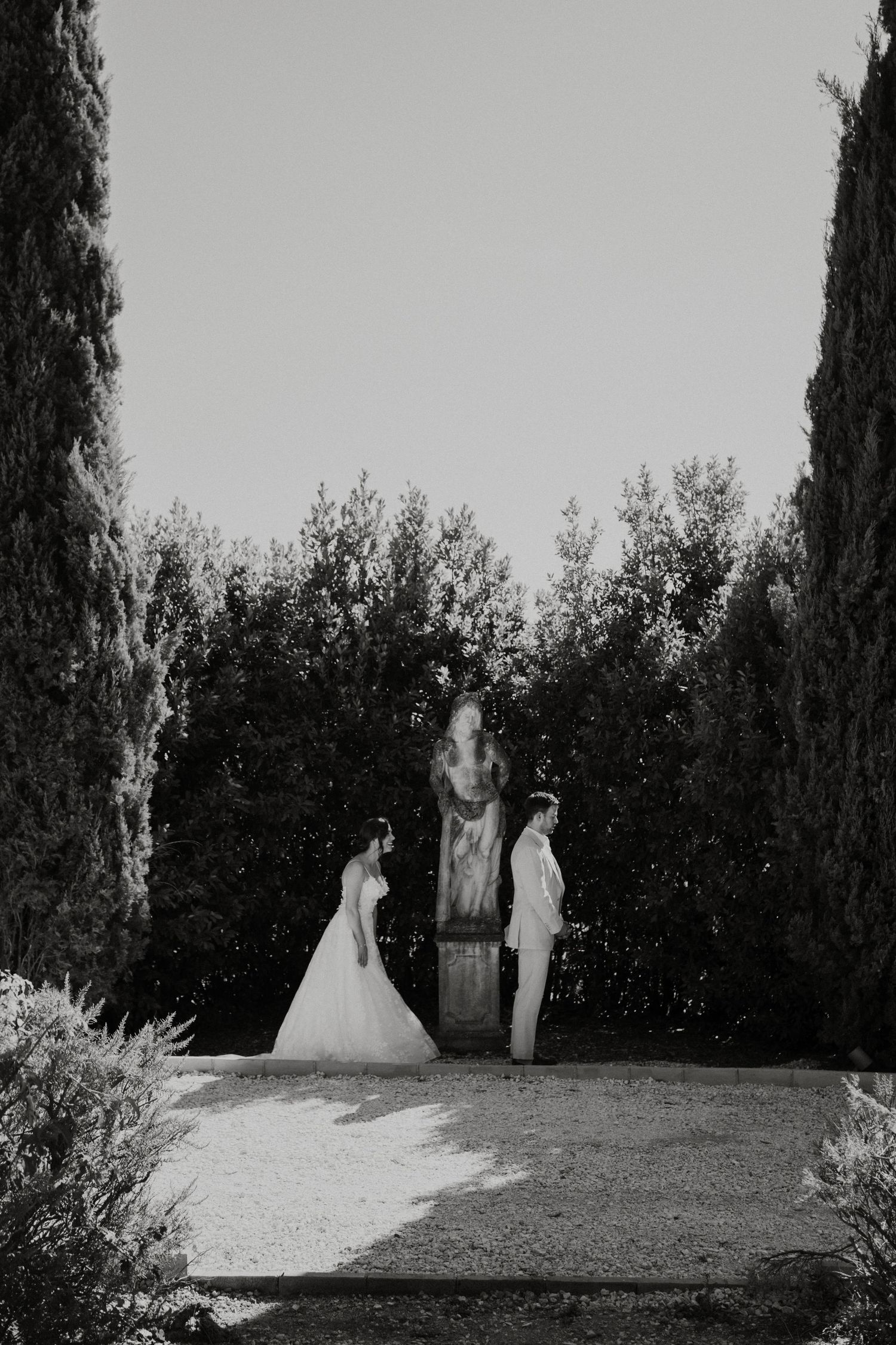 Black and white wedding photo of bride and groom standing before a garden statue, framed by tall cypress trees.