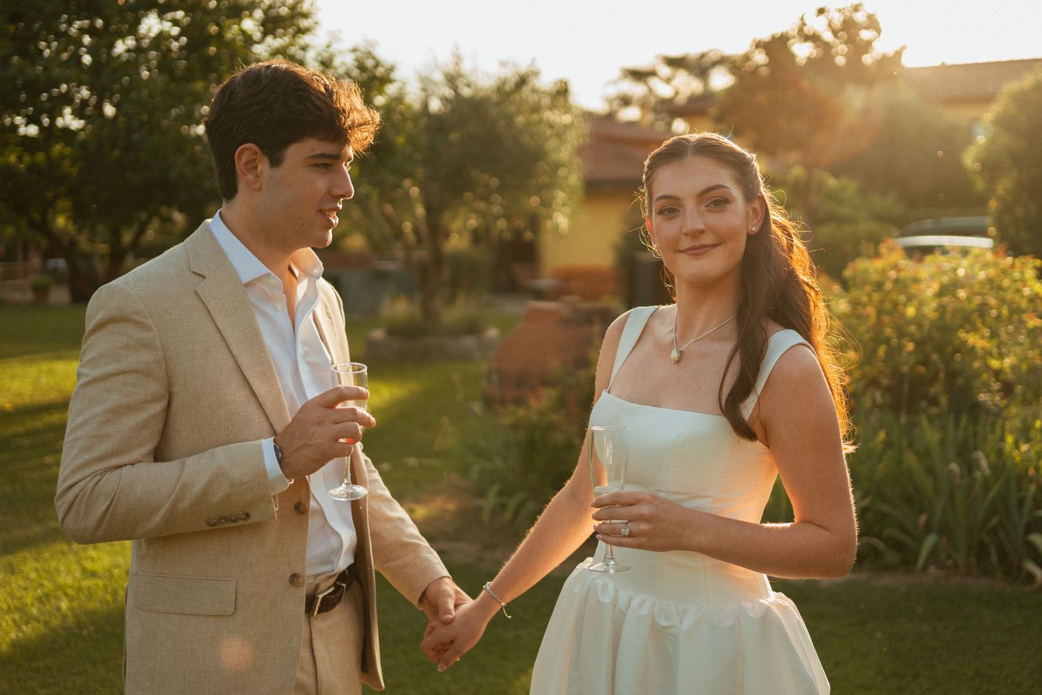Young couple holds champagne glasses and hands in sunlit garden, woman in white dress, man in beige suit.