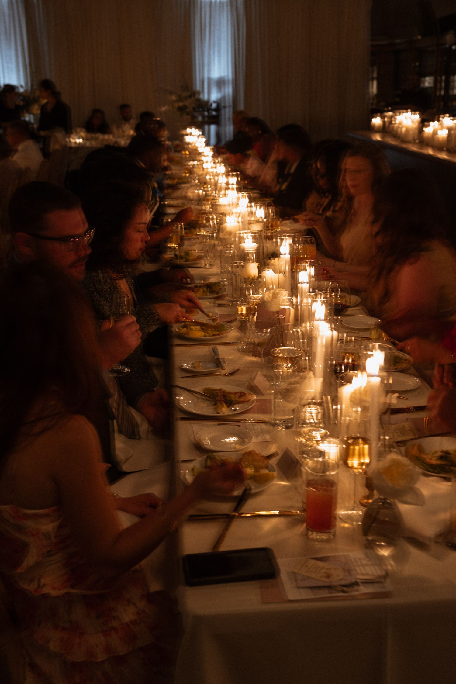 Wedding guests dining by candlelight at a long elegantly decorated reception table in dim lighting.