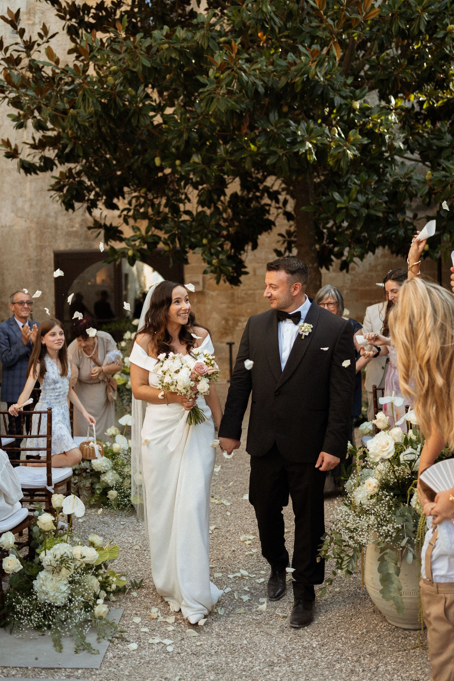 Bride and groom walk down outdoor aisle under magnolia tree at rustic Italian wedding venue.