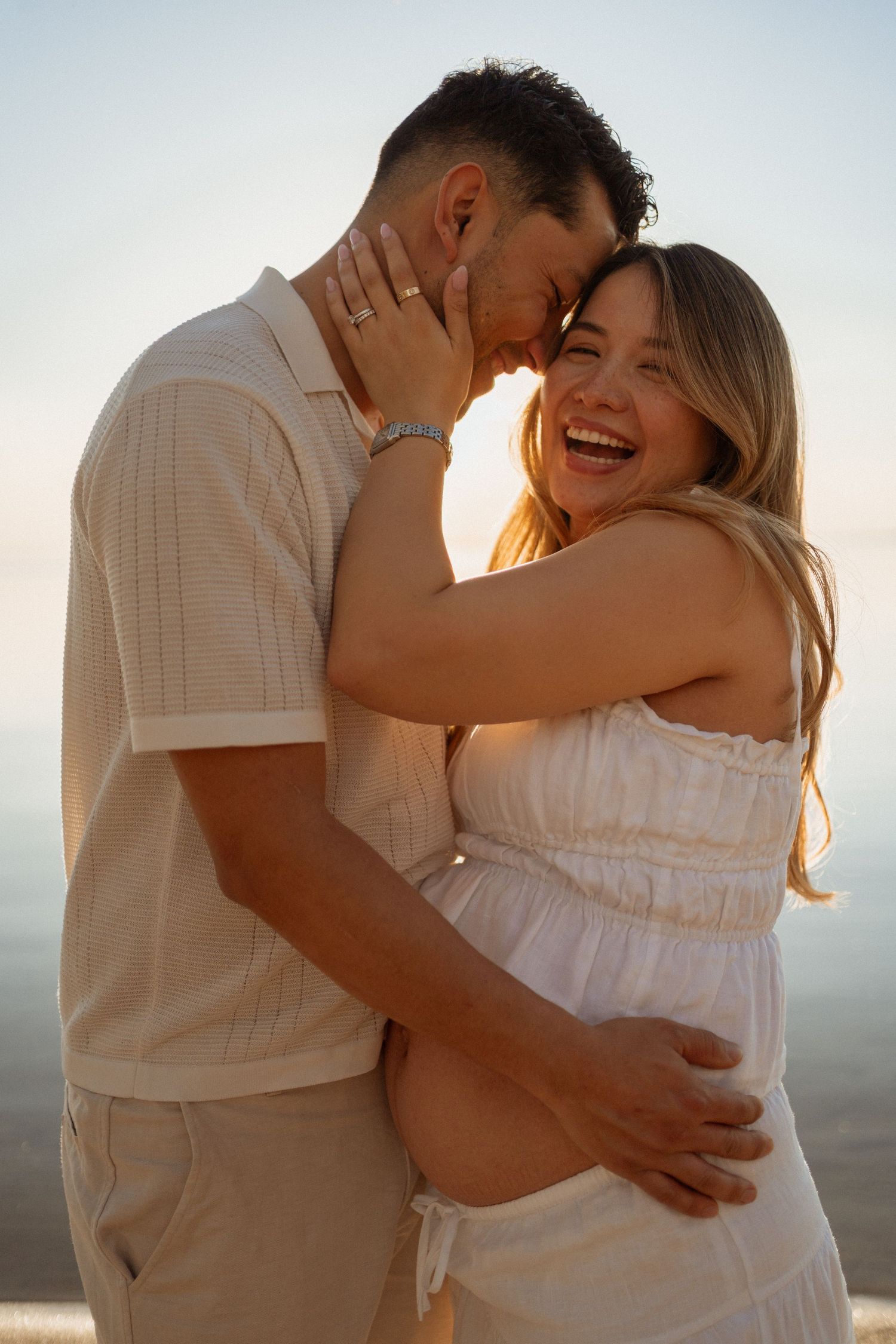 Happy pregnant couple embracing at golden sunset, woman laughing while man holds her baby bump.