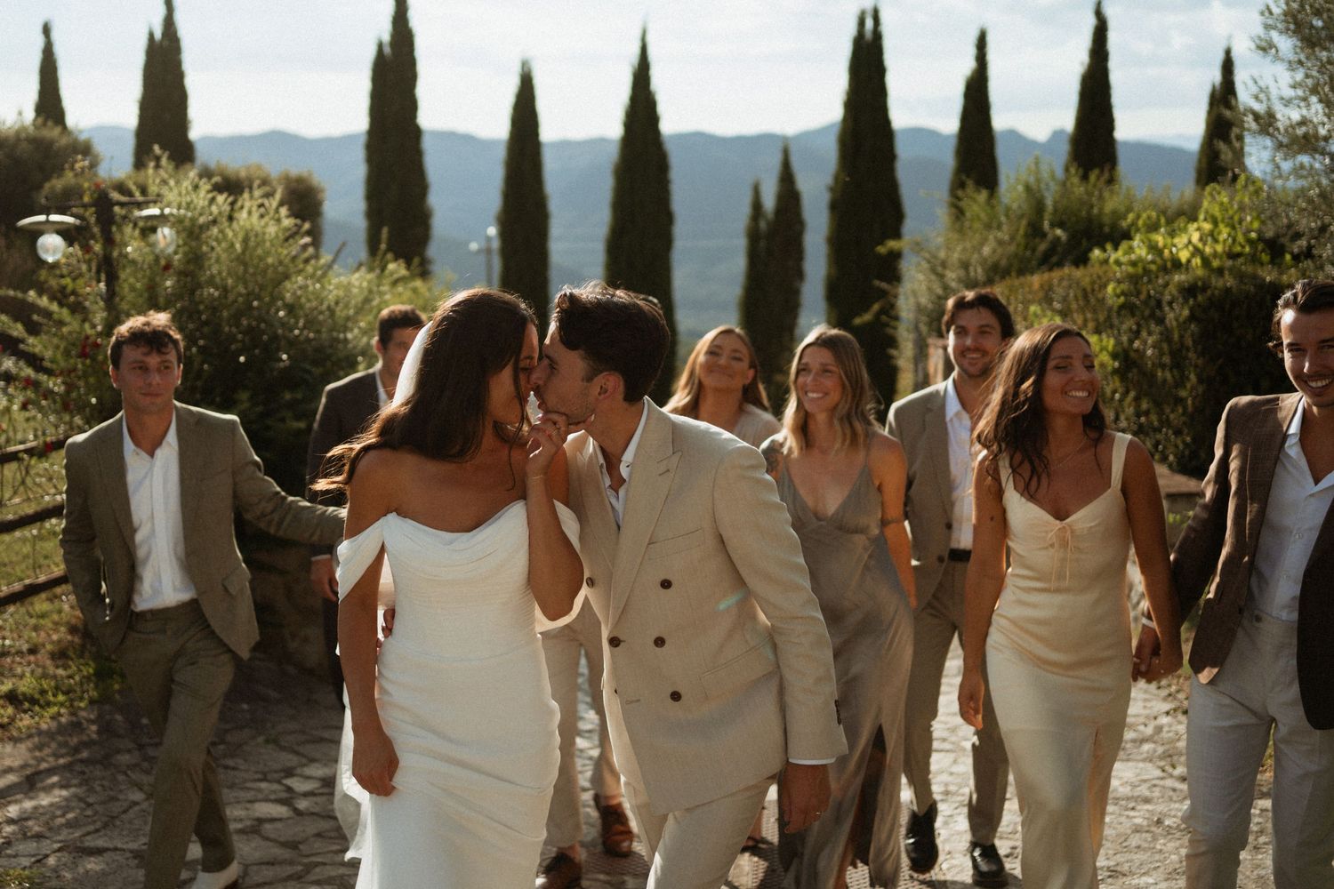 Bride and groom share a kiss while walking with wedding guests through a scenic Tuscan villa garden.