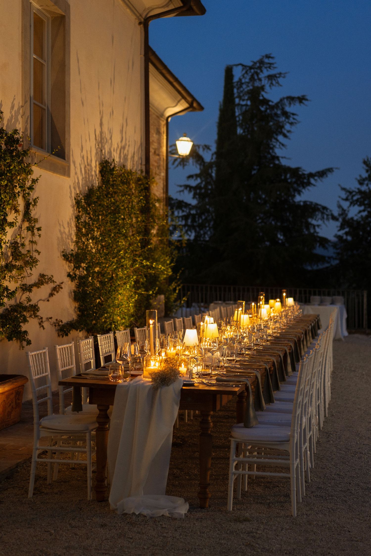Elegant outdoor candlelit dinner table set at dusk beside a Tuscan villa, with white chairs and glowing lanterns.