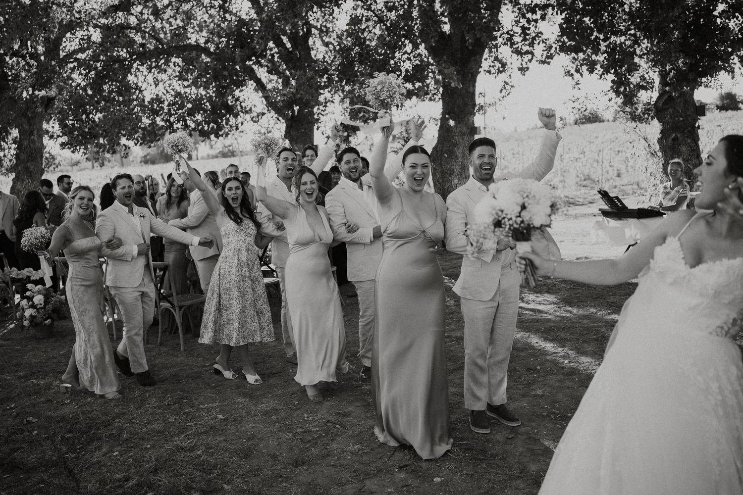 Black and white photo of wedding party celebrating outdoors under trees during a joyful ceremony reception.