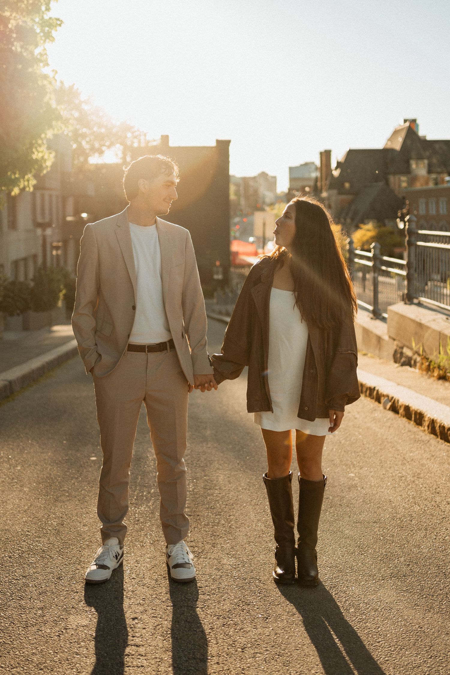 A couple holds hands while walking on a sunlit street, the man in a beige suit and the woman in a white dress with black boots.