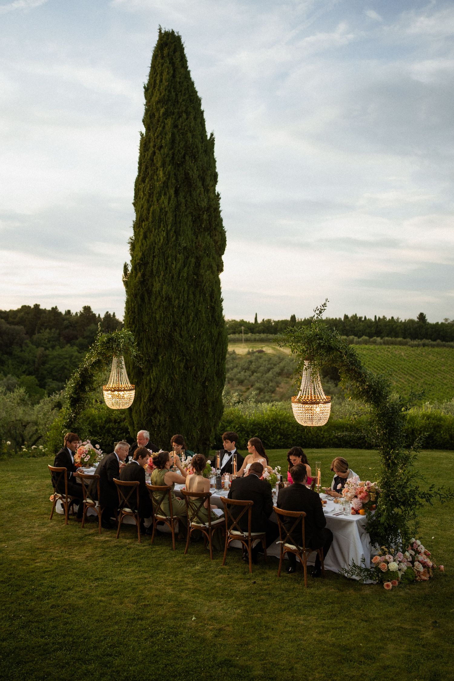Elegant outdoor wedding reception dinner under chandeliers beside tall cypress tree in scenic Tuscan countryside at dusk.